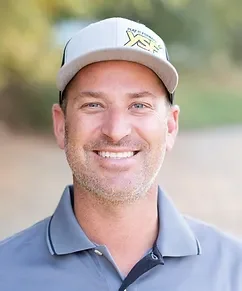 A smiling man wearing a white baseball cap and a blue collared shirt, outdoors with a blurred background of greenery.