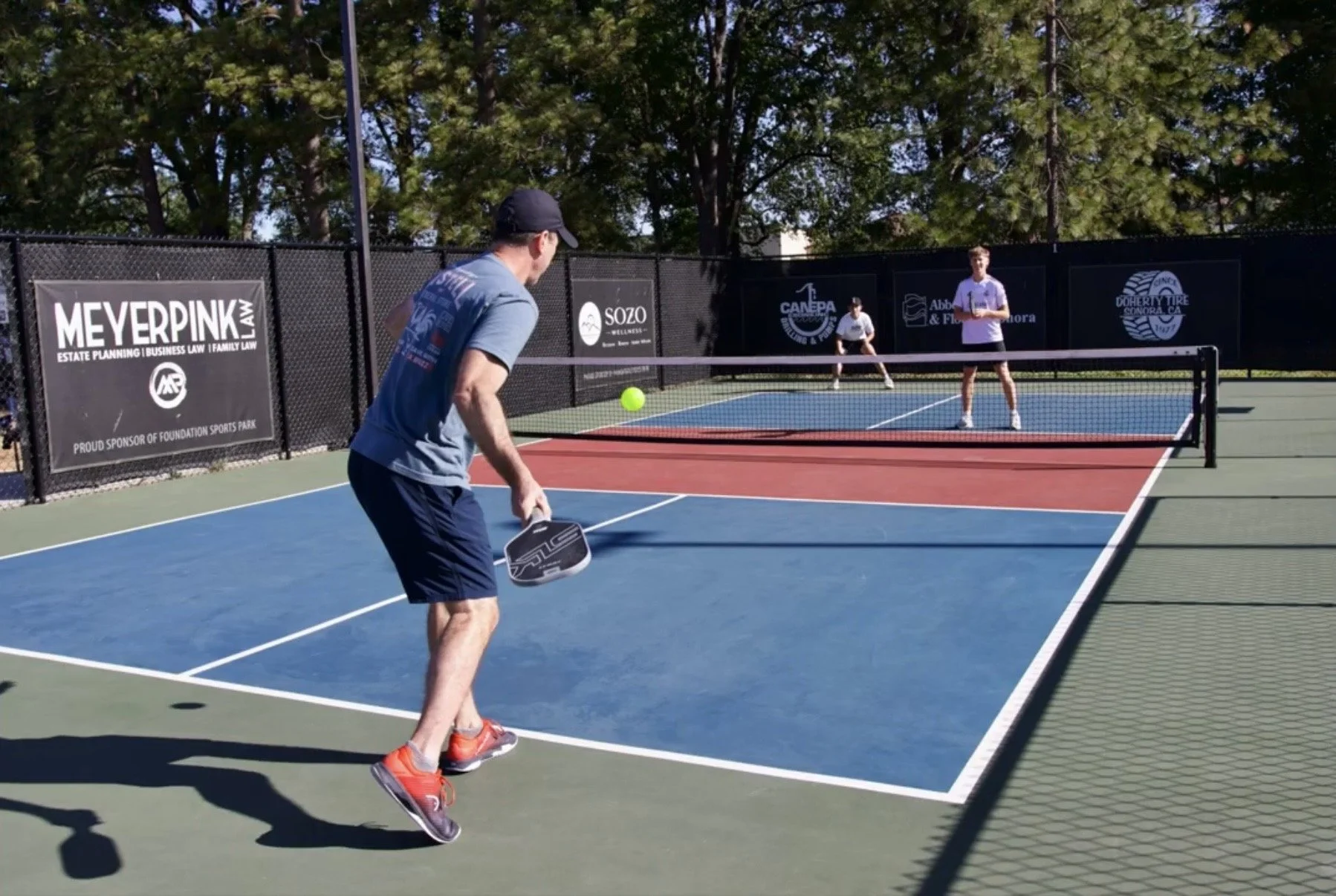 Three men playing pickleball on an outdoor court with trees in the background. One man in the foreground is about to hit the ball with a paddle, while two others stand on the opposite side of the net.