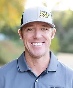 Portrait of a smiling man wearing a gray cap with a yellow logo and a gray polo shirt outdoors with blurred green trees in the background.