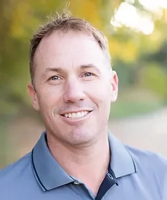 Close-up of a smiling man wearing a blue collared shirt, outdoors with blurred greenery in the background.