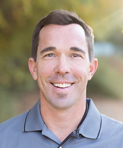 A smiling man outdoors with a blurred green and yellow background, wearing a gray collared shirt.