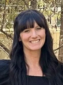 A woman with long dark hair and bangs smiling outdoors against a background of trees and foliage.