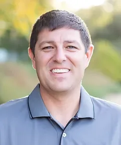 A smiling man with short dark hair wearing a blue collared shirt outdoors with greenery in the background.