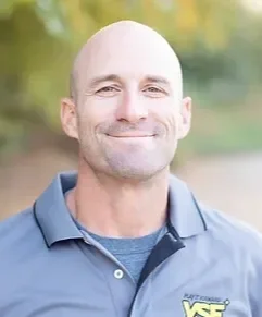 A smiling bald man wearing a blue collared shirt with a logo outdoors