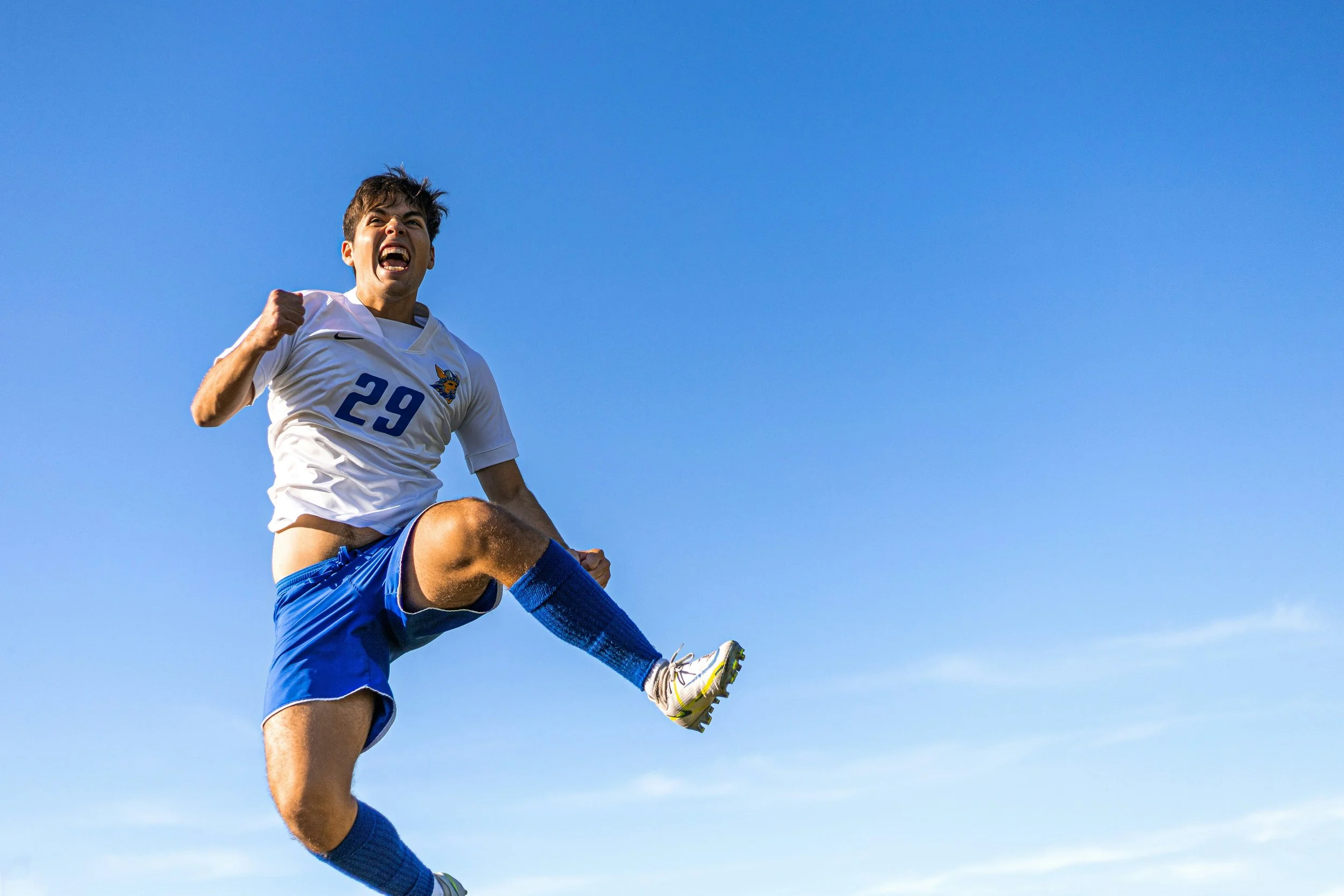 A young male soccer player in a white jersey with the number 29 and a lion logo, blue shorts, and blue socks, celebrating with a fist pump while jumping in the air against a clear blue sky.