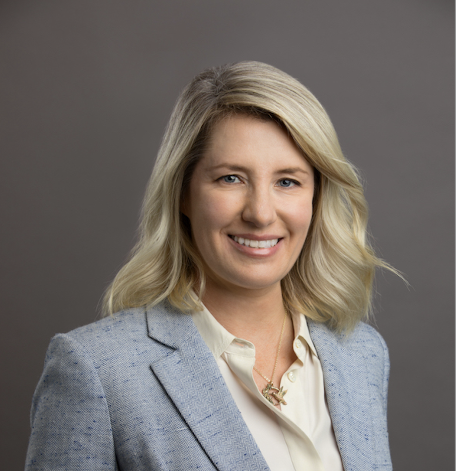 A professional woman with blonde hair and a smile, wearing a light blue blazer and a cream blouse, against a gray background.