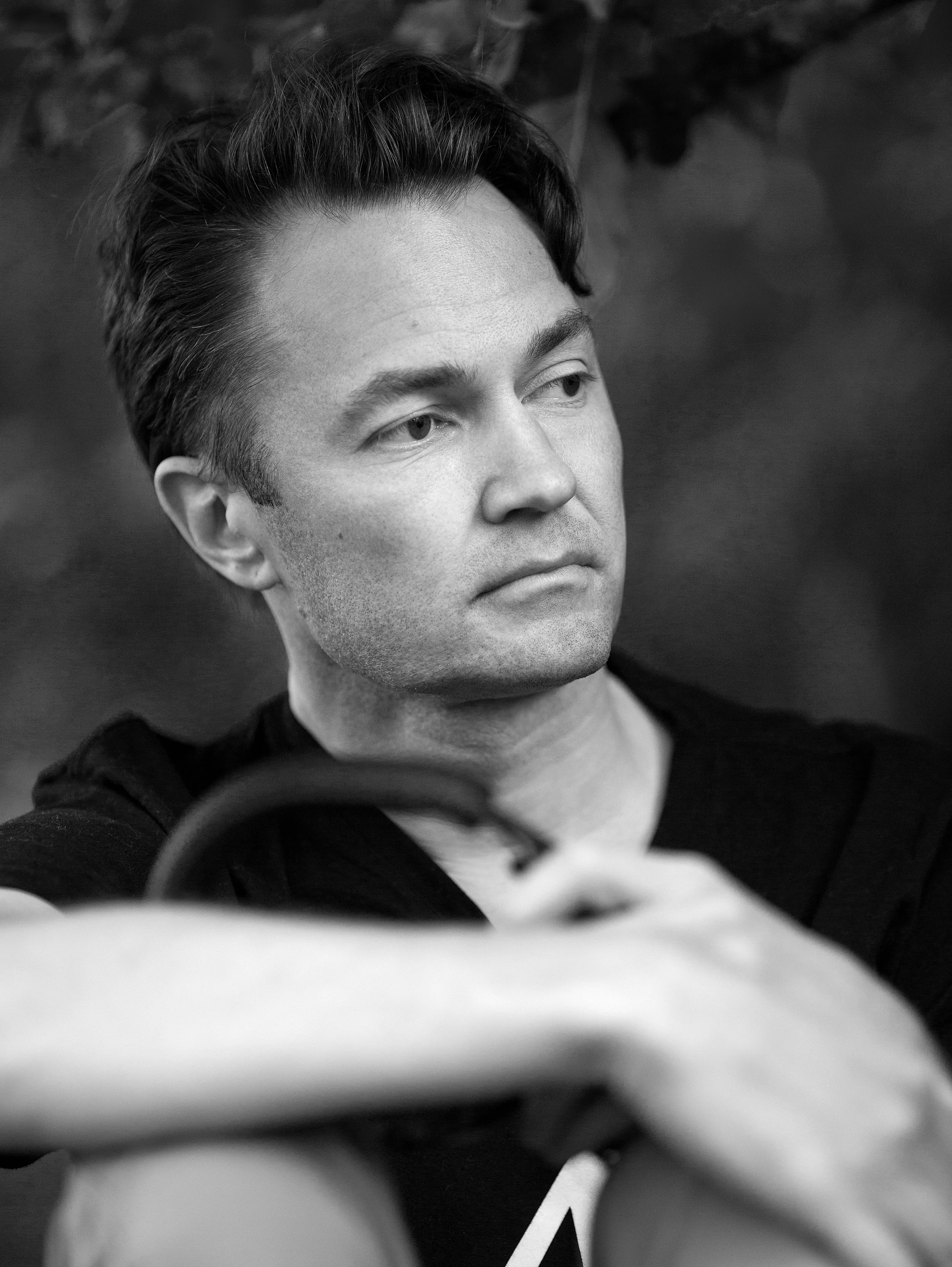 Close-up black and white photo of a young man with styled hair, looking slightly to the side. His face is in focus, with a blurred background.