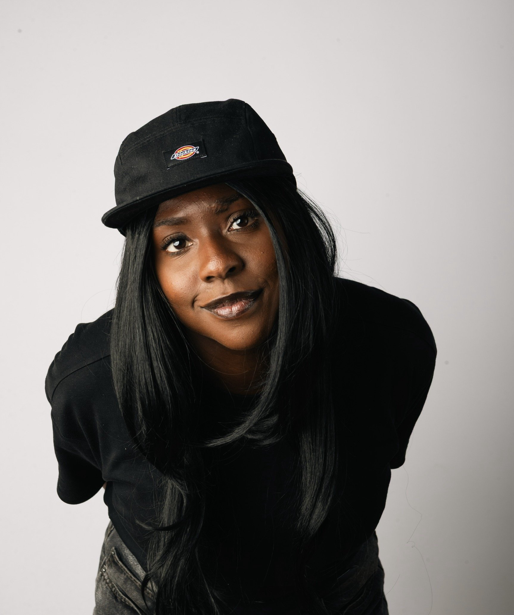 A woman with long black hair wearing a black Dickies cap and black shirt, smiling slightly against a plain gray background. DJ Savannah drum and bass DJ press shot.