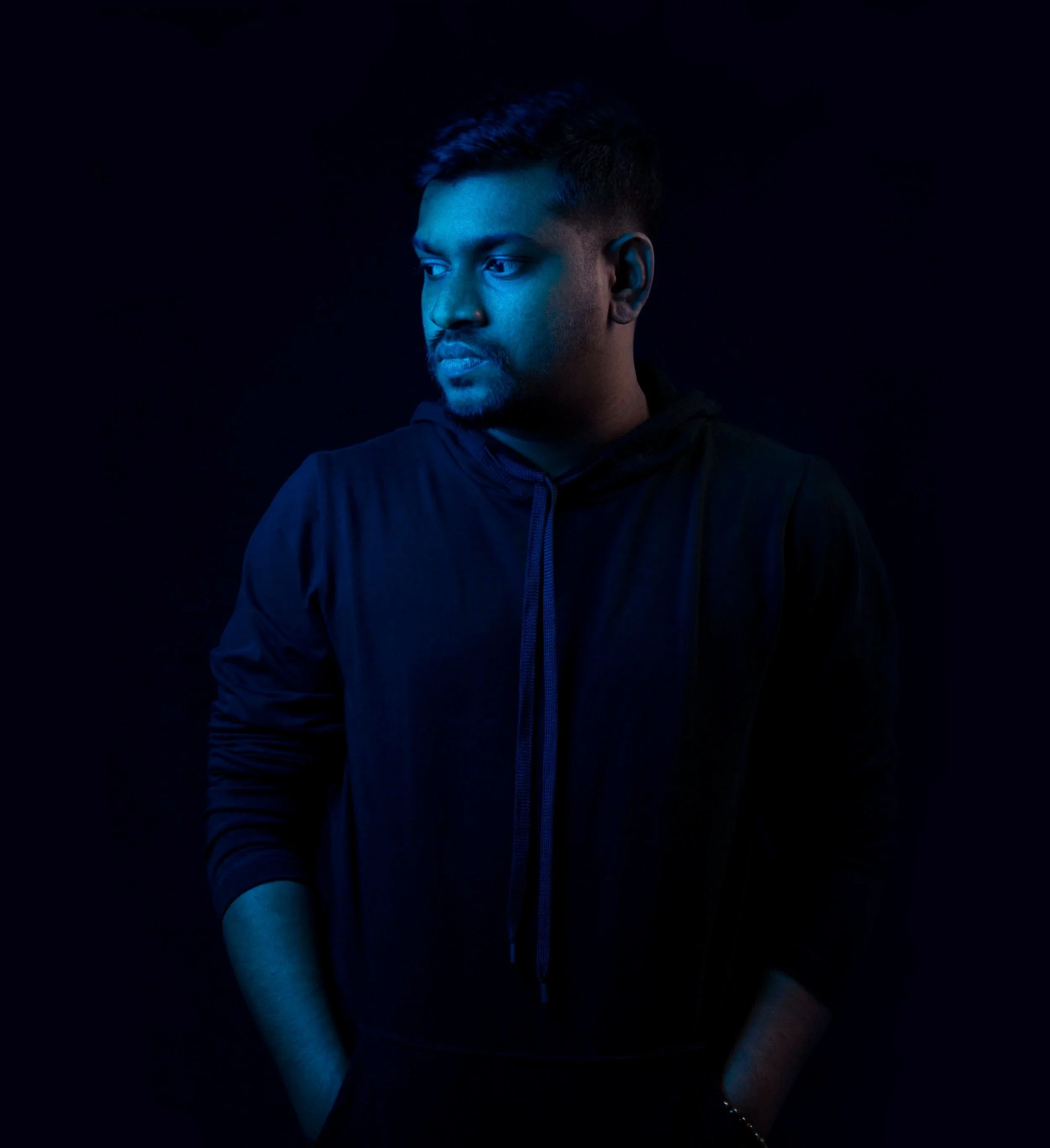 A man with short hair and a beard looking to the left, illuminated by blue lighting in a dark setting, wearing a black hoodie. DJ IYRE press shot.