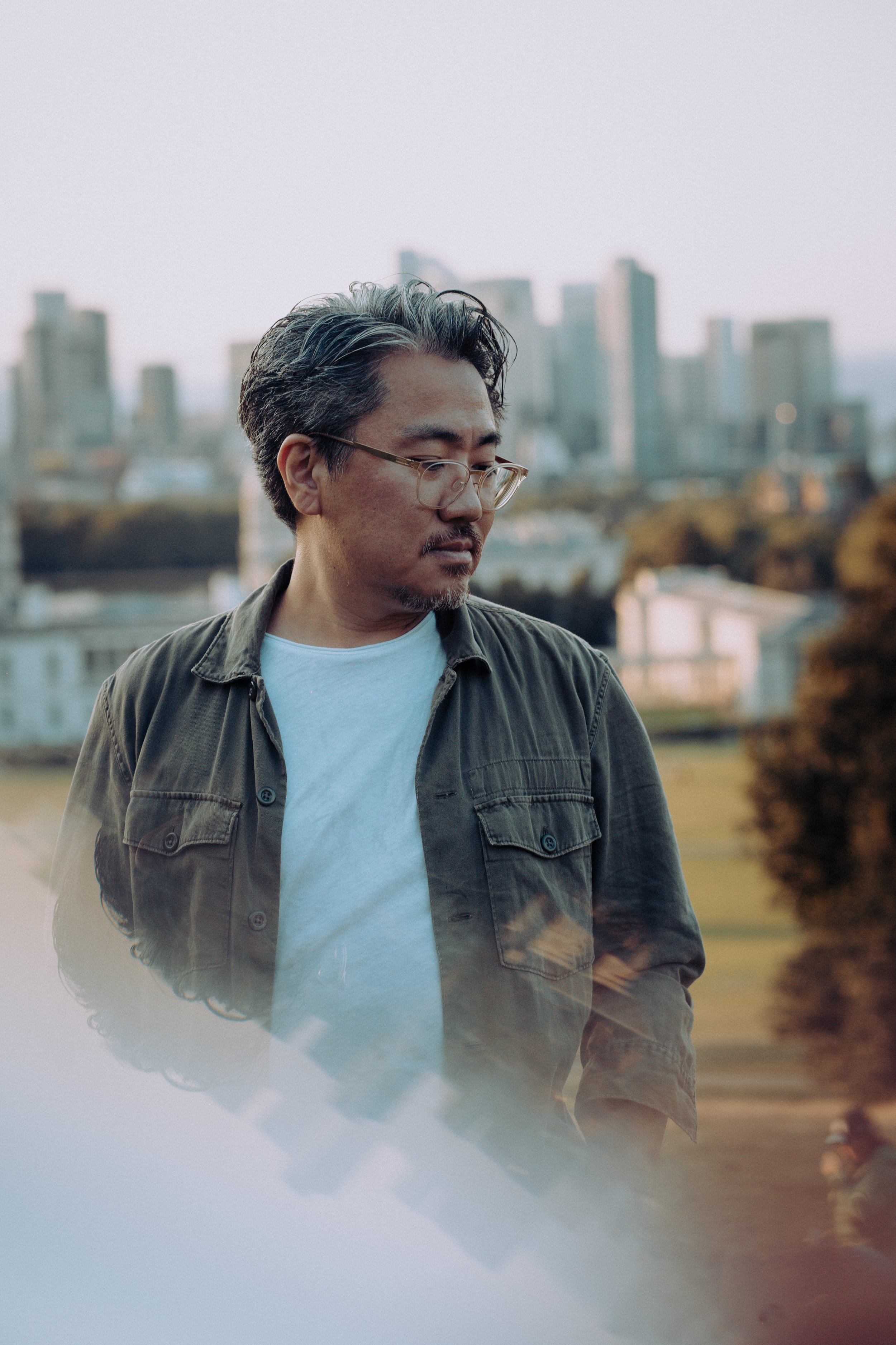 A man with glasses and a mustache wearing a brown jacket and white t-shirt stands outdoors with a city skyline in the distance. Makoto drum and bass DJ press shot.