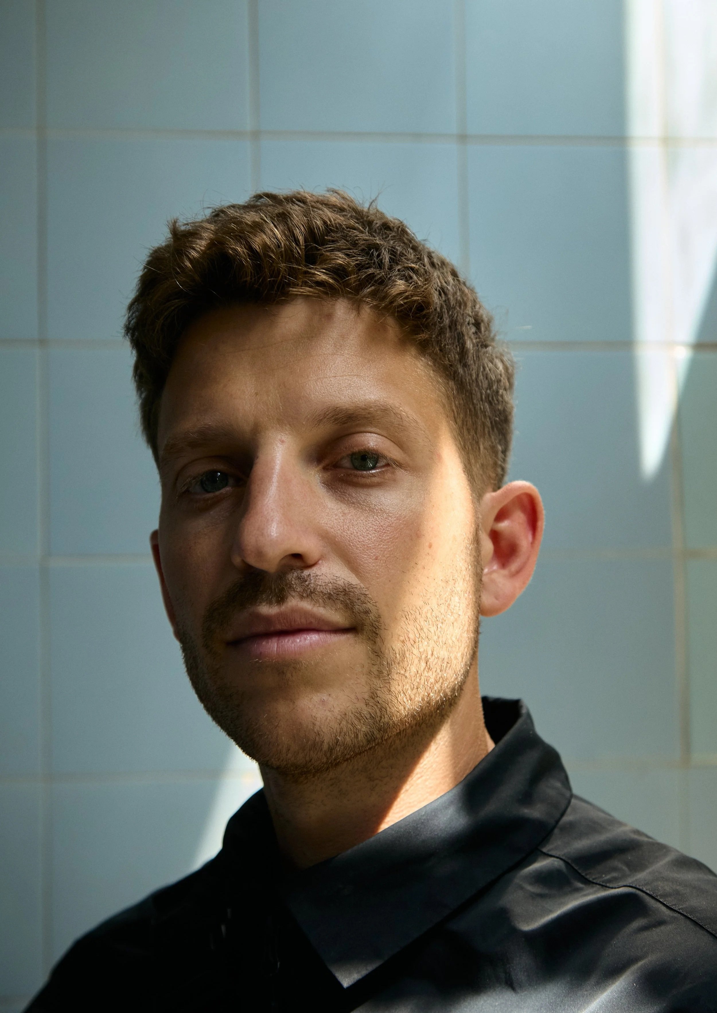 A young man with short brown hair, blue eyes, and light facial hair, wearing a black jacket, standing in front of a tiled wall with sunlight shining on his face. Hugh Hardie drum and bass DJ press shot.
