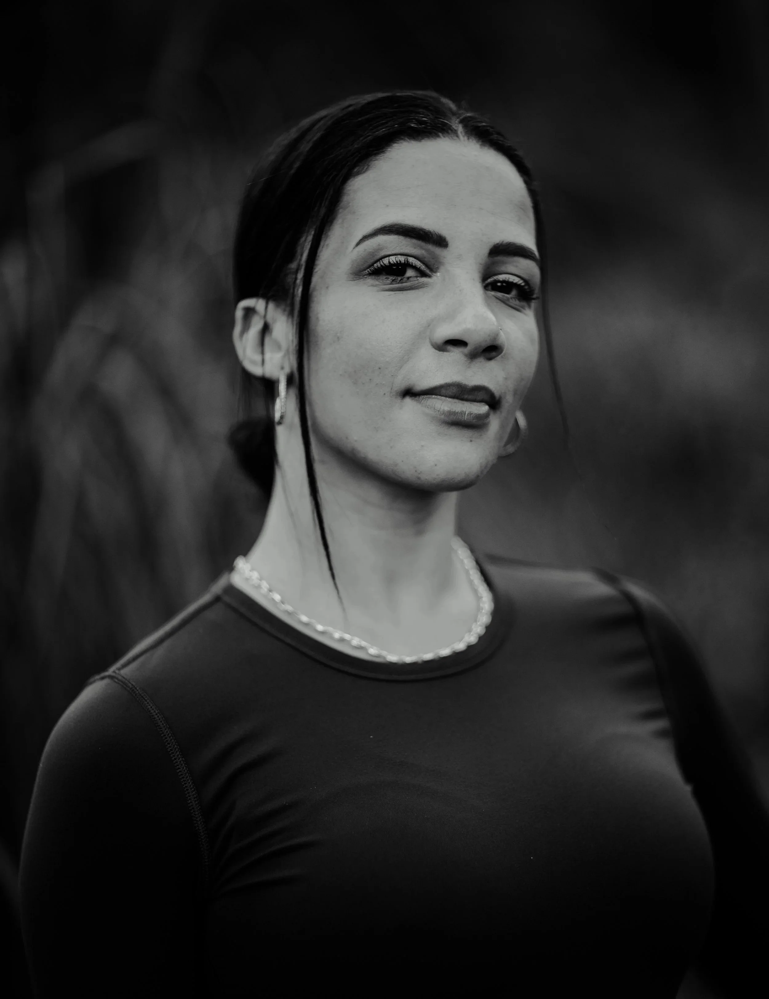 Black and white photo of a woman with dark hair styled back, wearing a dark top, pearl necklace, earrings, and makeup, with a confident expression. Drum and bass DJ Stay-C press shot.