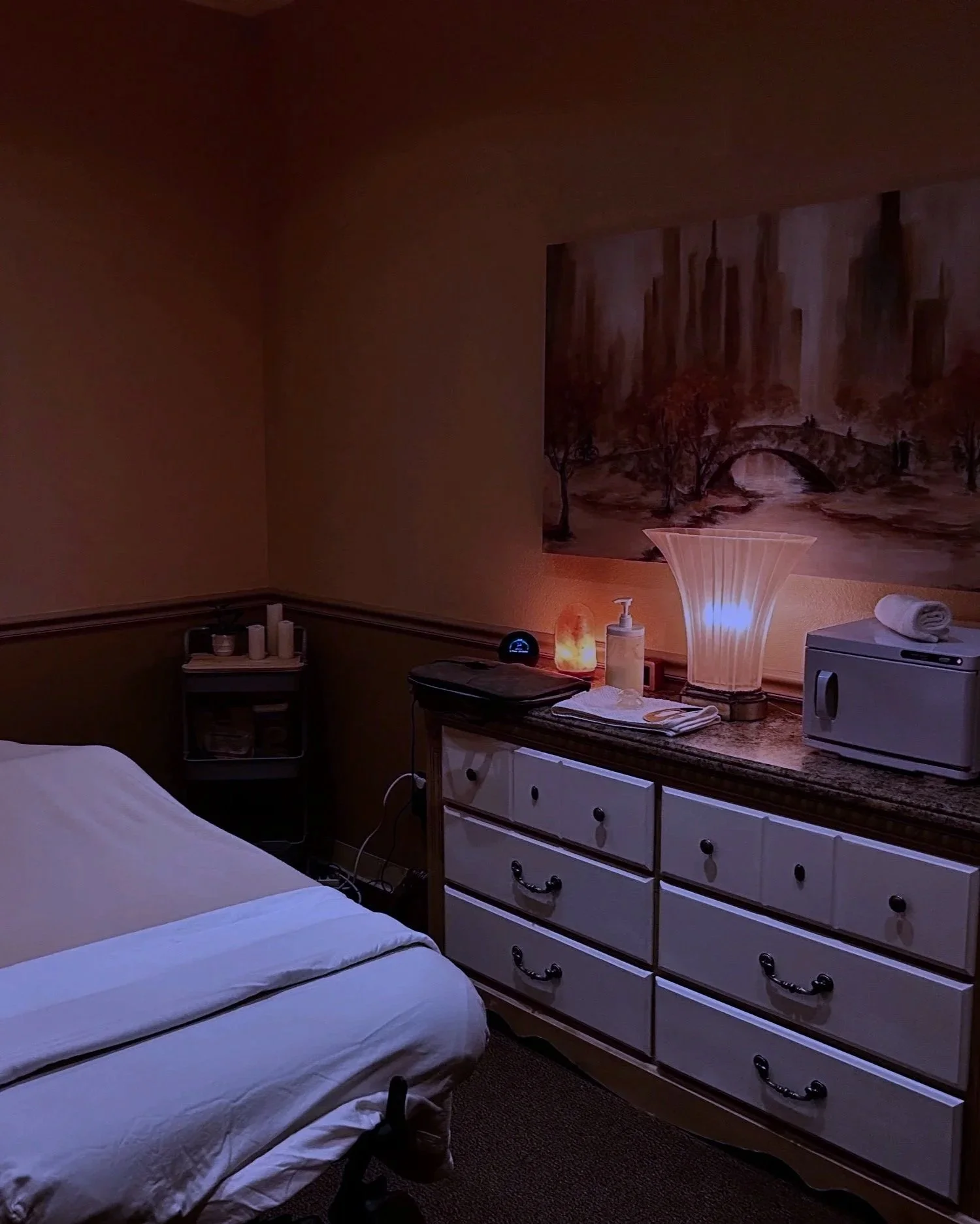 Dimly lit bedroom with a dresser and a painting on the wall, candles and decorative items on top, a lamp emitting warm light, a folded towel on a microwave, and part of a bed with white bedding.
