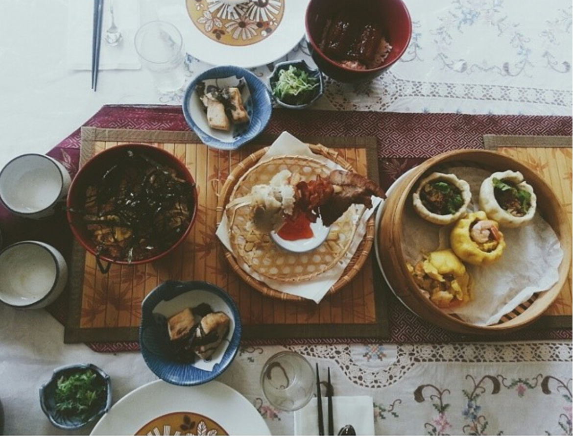 A table with a steamer of homemade open faced baos, bowls of unagi don, a small hanger with sliced meal draped off of it, two small bowls with white fish, and small side dishes with seaweed salad