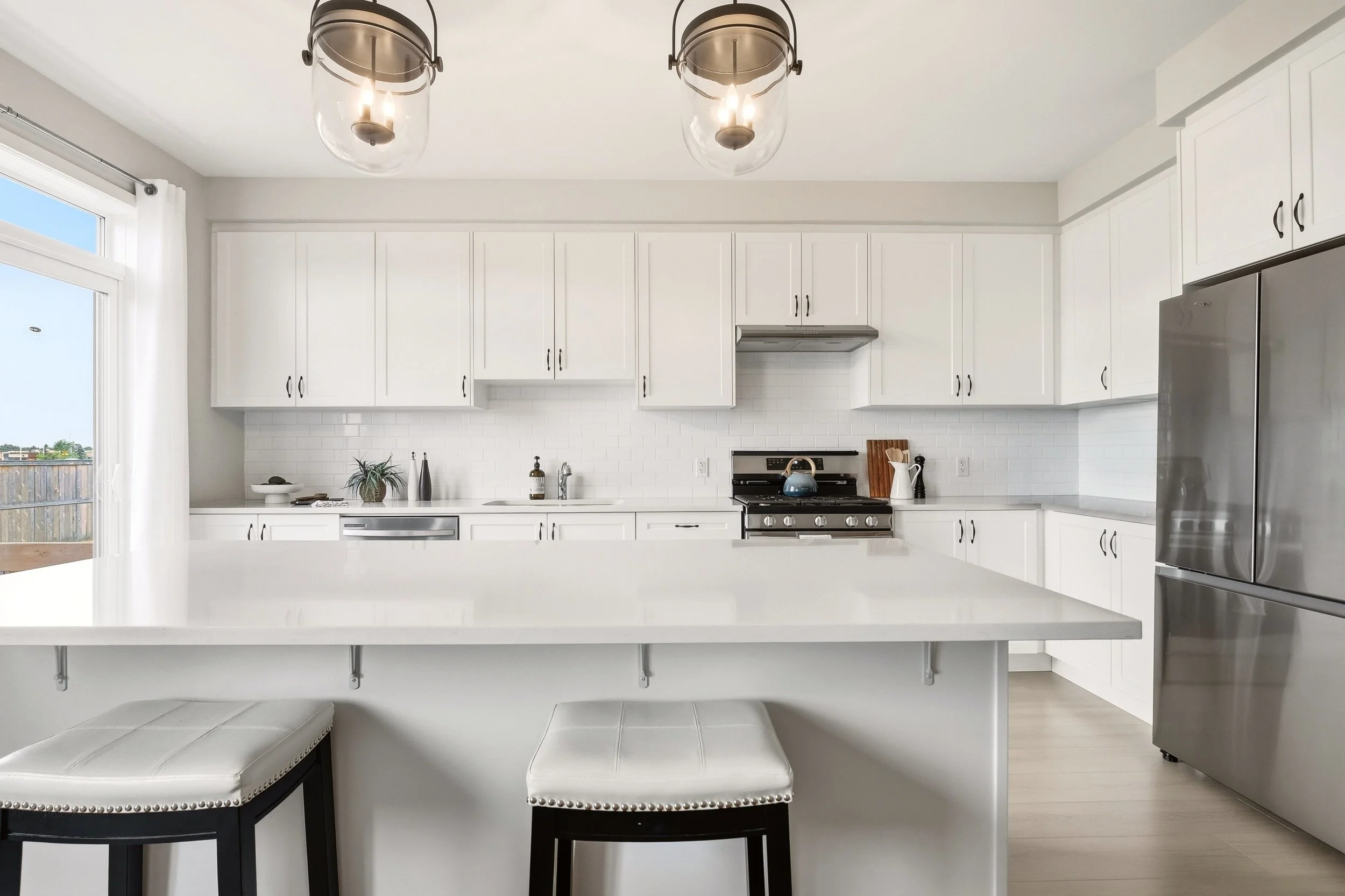 Modern white kitchen with pendant lights, stainless steel appliances, and a breakfast bar with two white padded stools.