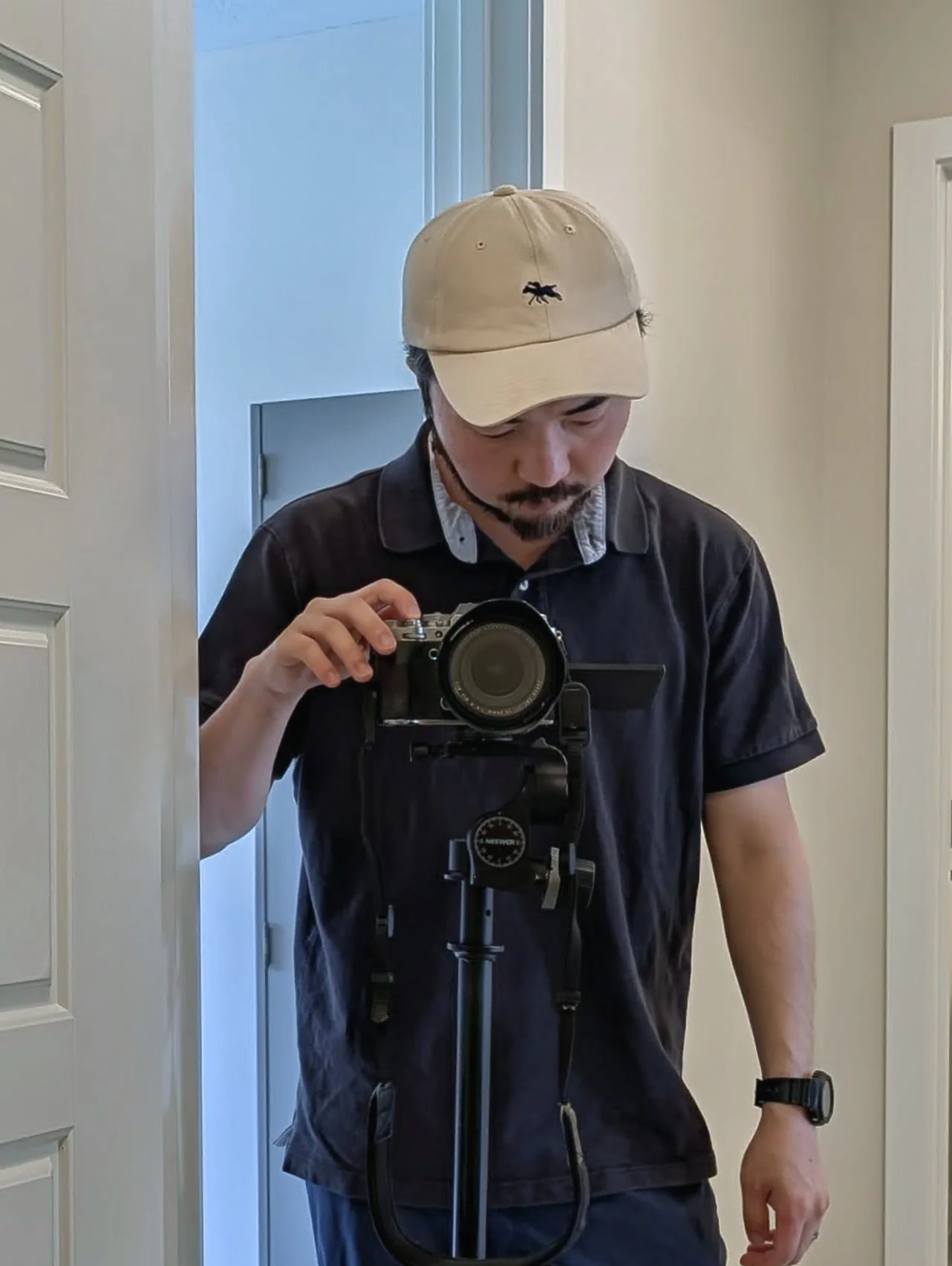 A man wearing a beige baseball cap and black shirt is taking a photo of himself in a mirror with a camera mounted on a tripod. The man is looking down at the camera and standing near a white door with a white wall background.
