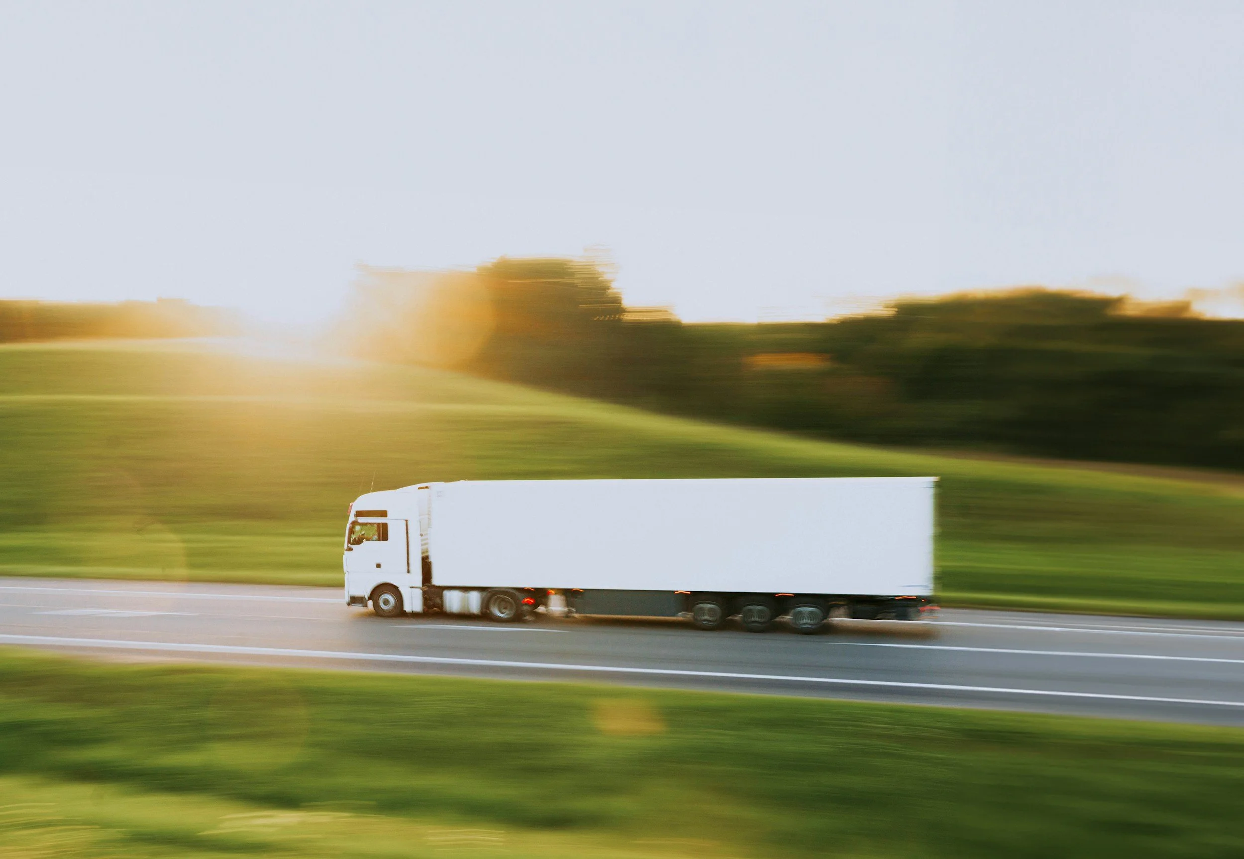 A white semi-truck with a trailer driving on a highway with a green grassy landscape and trees in the background during daylight.