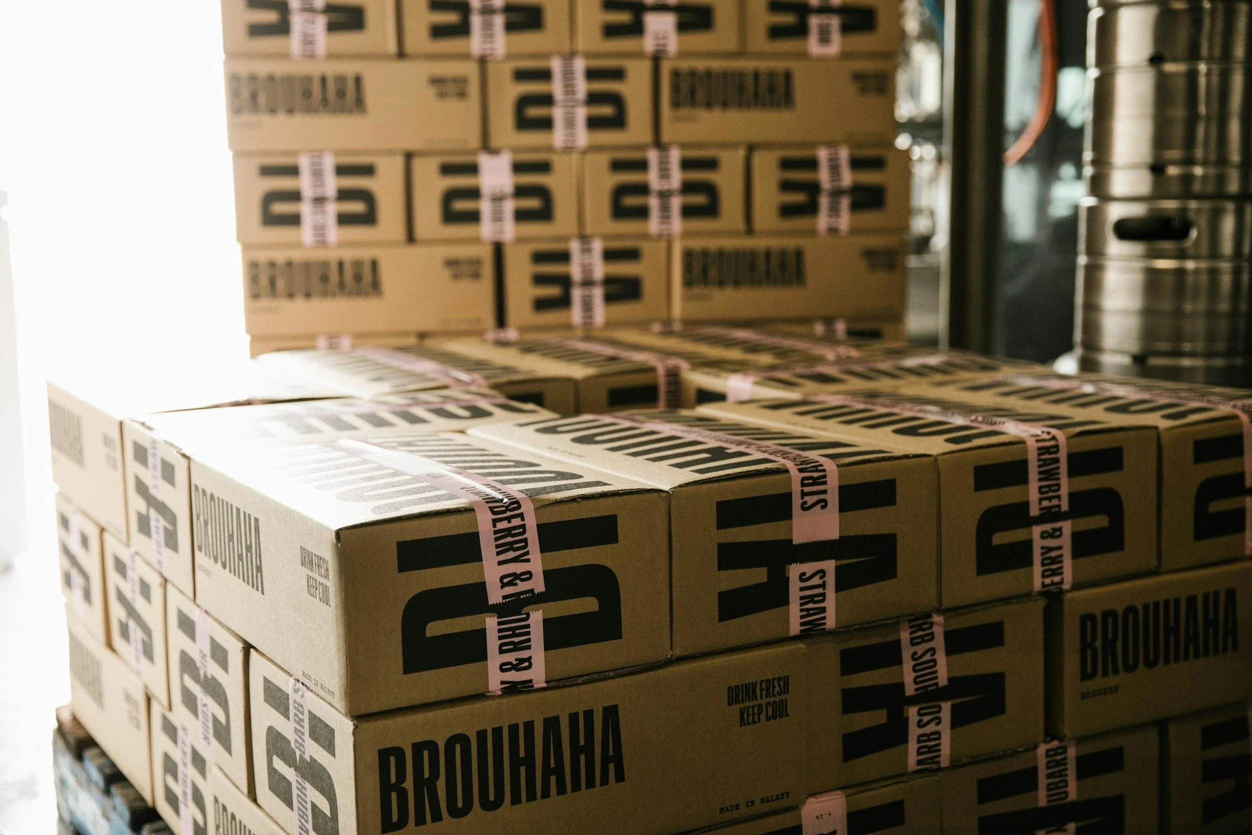 Boxes stacked on top of each other with labels that say "Brohahaha" and pink tapes labeled "Drink Fresh Keep Cool" in a storage or warehouse setting.