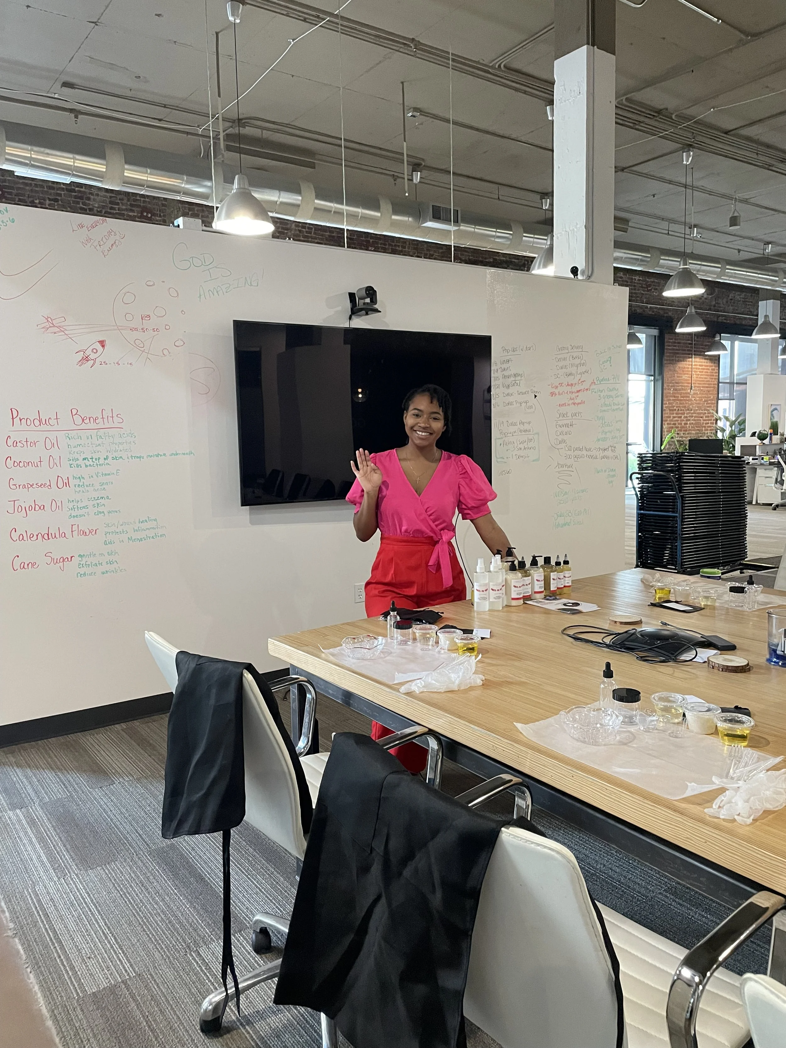 A woman in a pink shirt and red apron standing in an office or classroom, smiling and waving. Behind her, a whiteboard with notes and drawings, and a large flat screen monitor. The table in front has bottles, small jars, and scientific or cosmetic supplies. There are chairs with black and white covers and a stack of black chairs on the right. The room has exposed brick walls, large windows, and industrial ceiling lights.