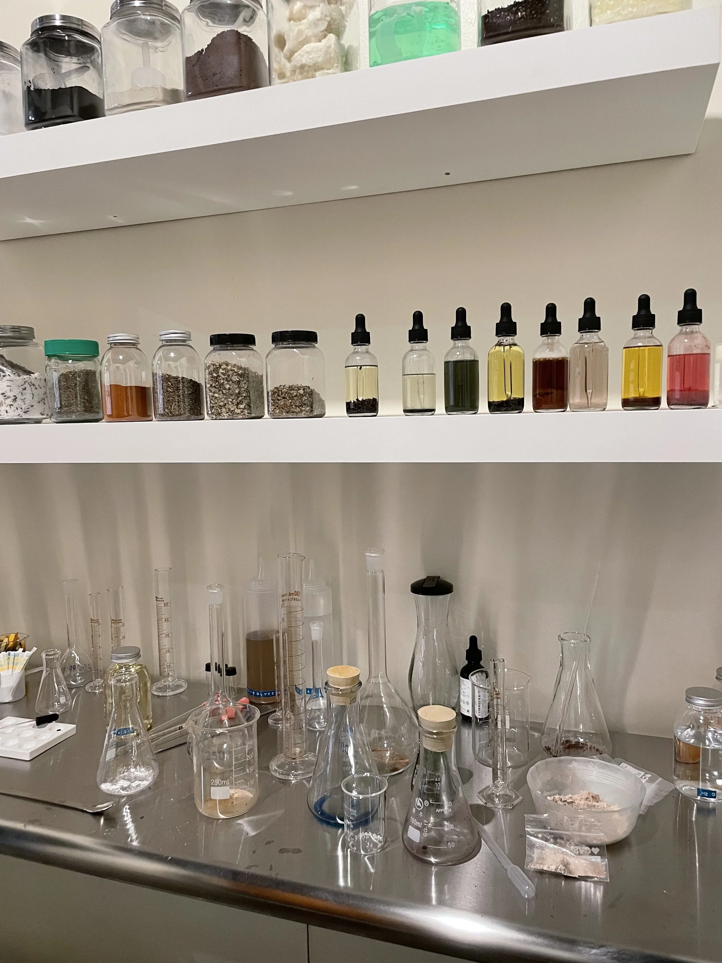 A laboratory workspace with various glass beakers, test tubes, and bottles containing different liquids, powders, and materials, some with droppers. Shelves above hold jars with powders and chemicals.