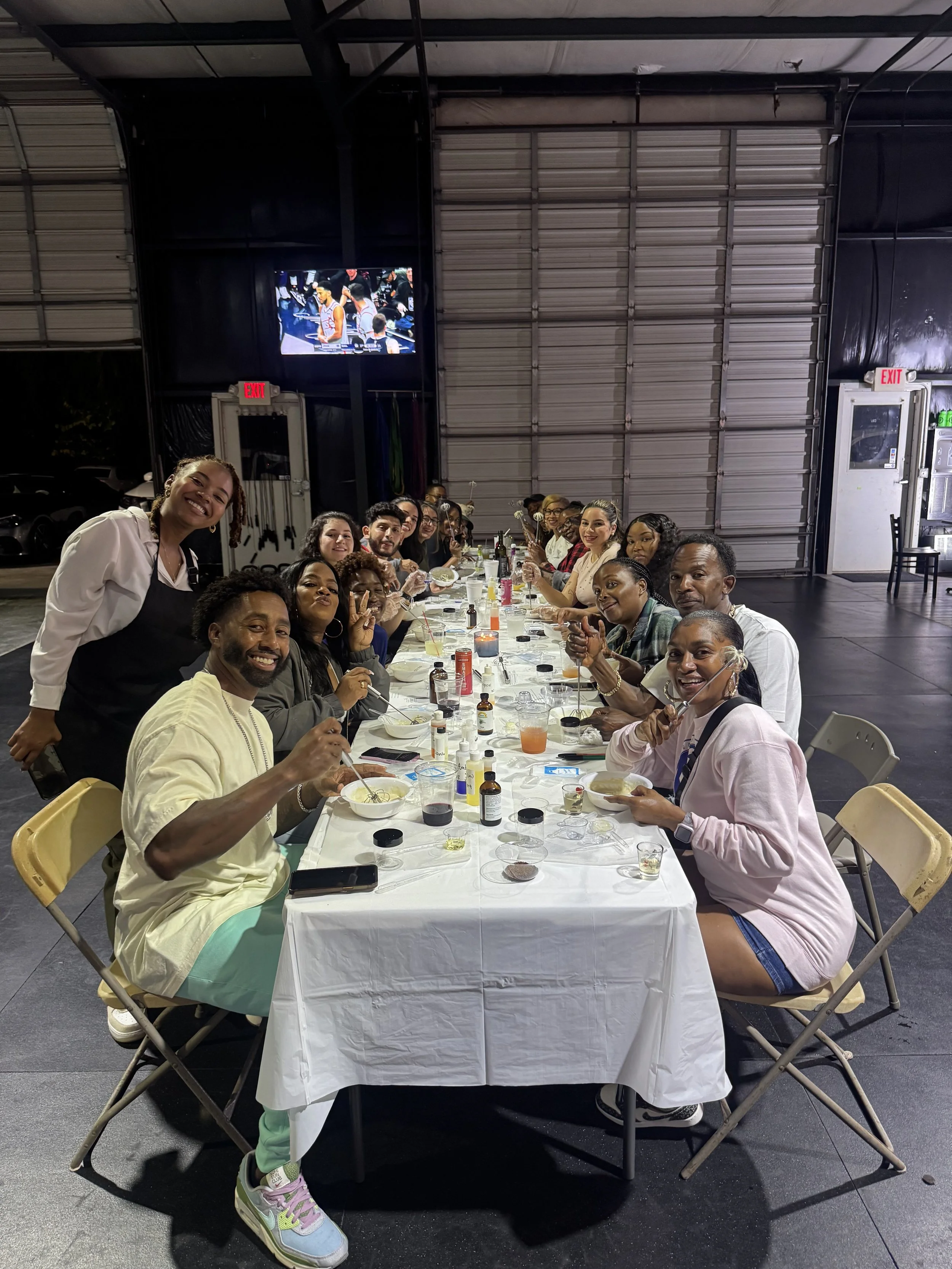 A large group of people are sitting around a long table at a gathering or celebration, with some holding forks and bowls of food, in an indoor space with a high ceiling and a metal garage door.