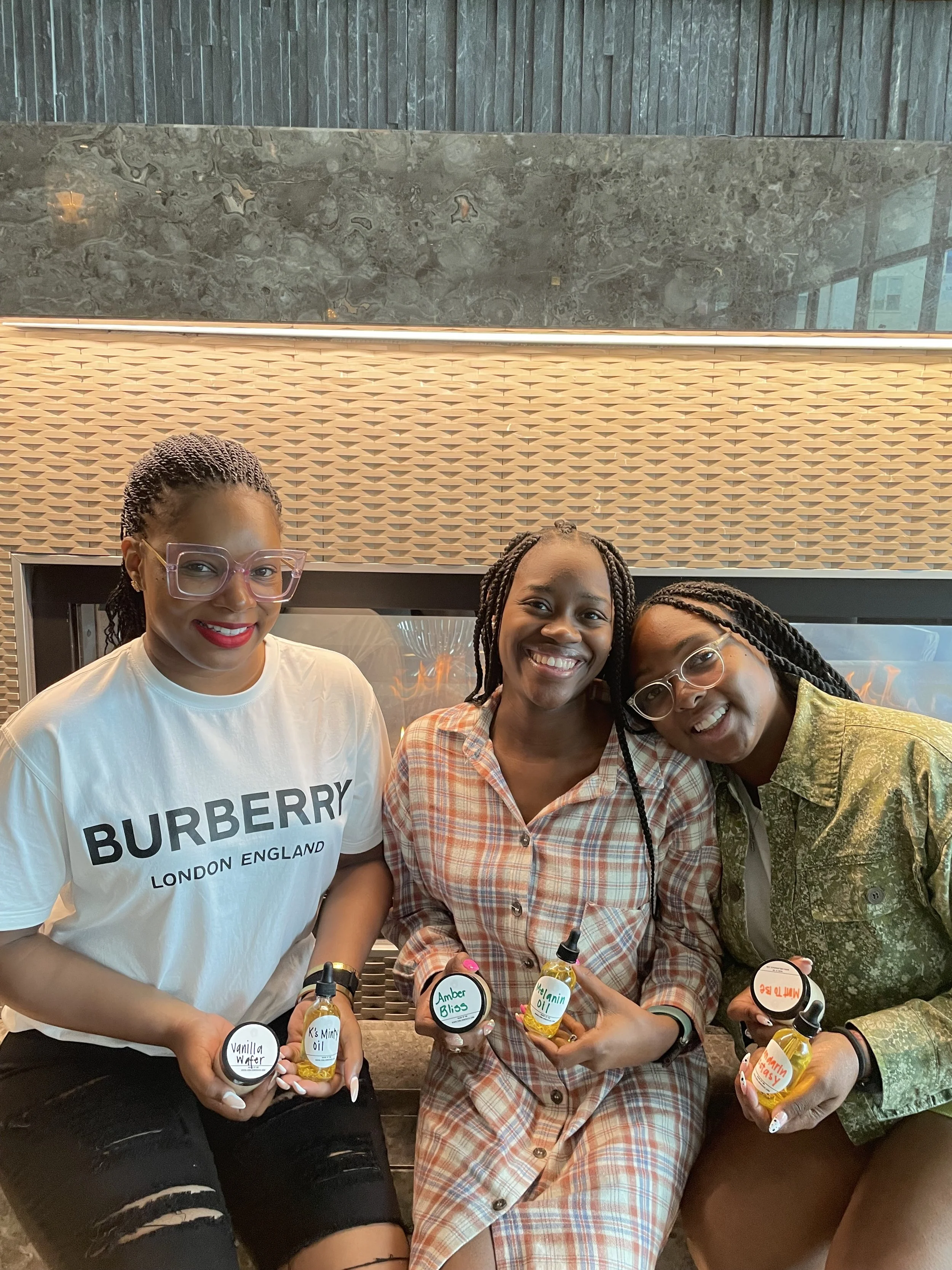 Three women sitting together, smiling and holding small bottles of essential oils labeled with various scents, in front of a modern fireplace and decorative wall.