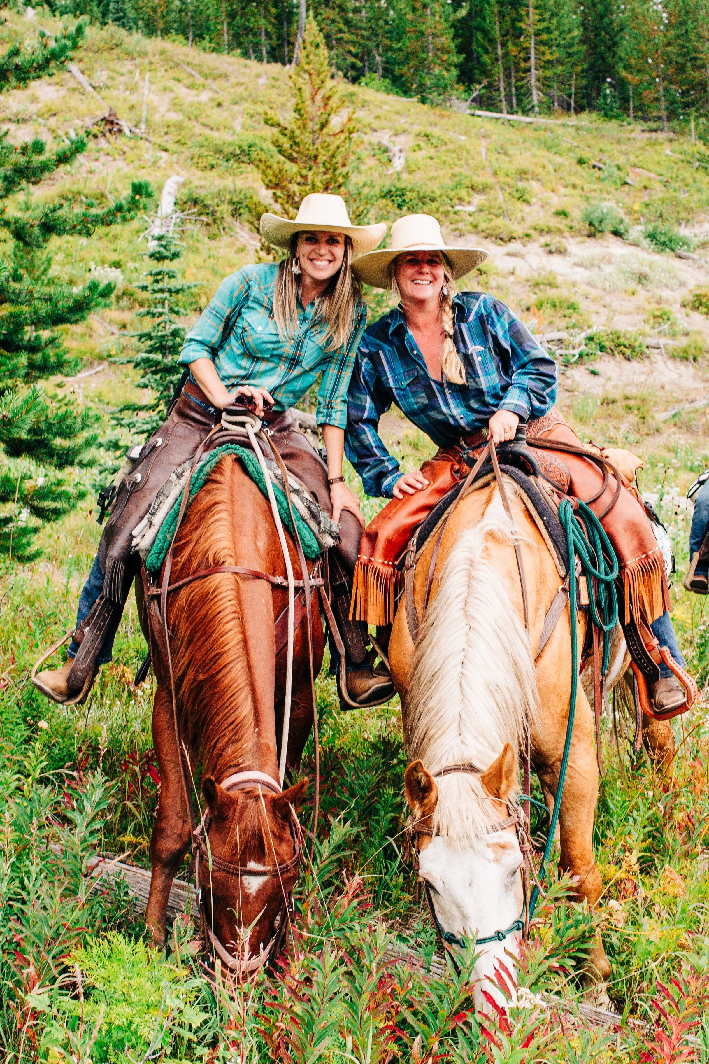 Two women riding horses in a lush green forest, wearing plaid shirts and large cowboy hats, smiling at the camera.