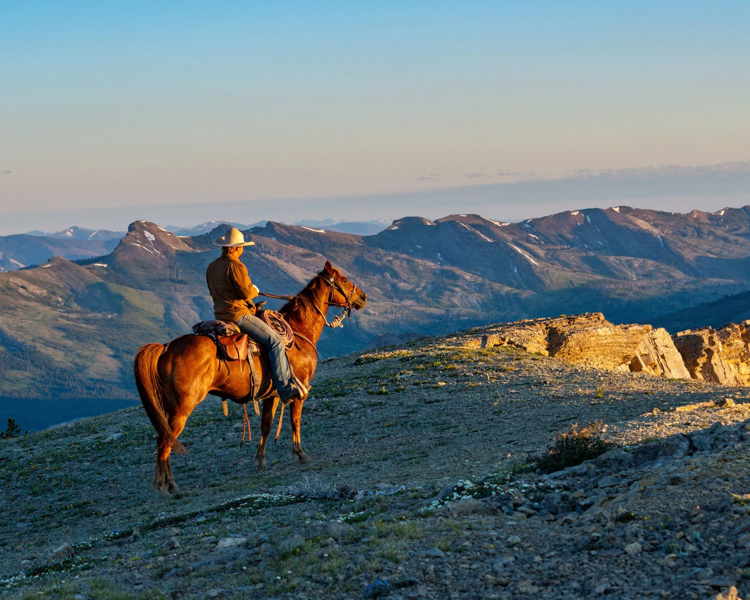 A person riding a horse in a mountainous landscape during sunset or sunrise, with snow-capped peaks in the background.