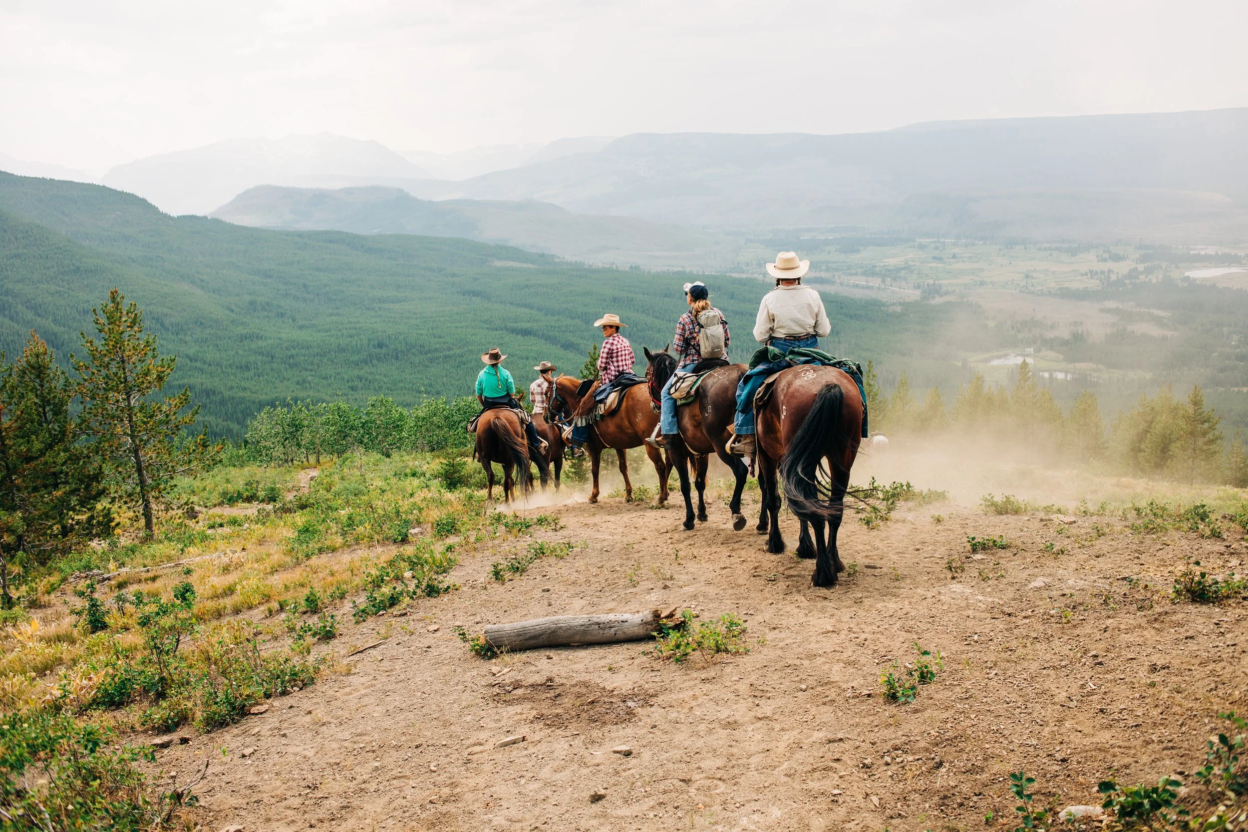 Group of people riding horses on a trail in a mountainous landscape with green forests and distant hills under a cloudy sky.