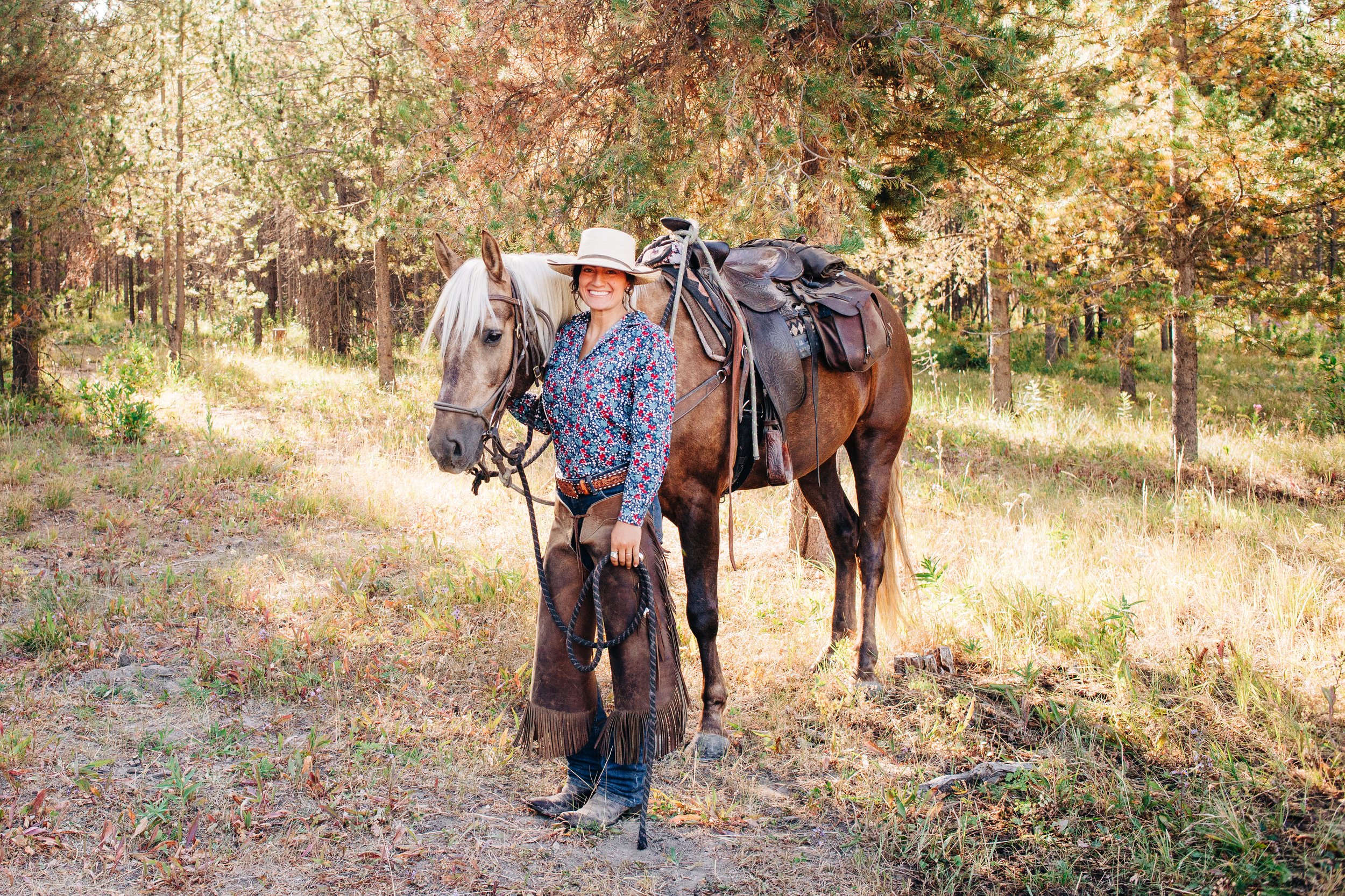 A woman with a big smile, wearing a hat and a floral shirt, standing beside a brown horse with a saddle, in a forested area with trees and grass.