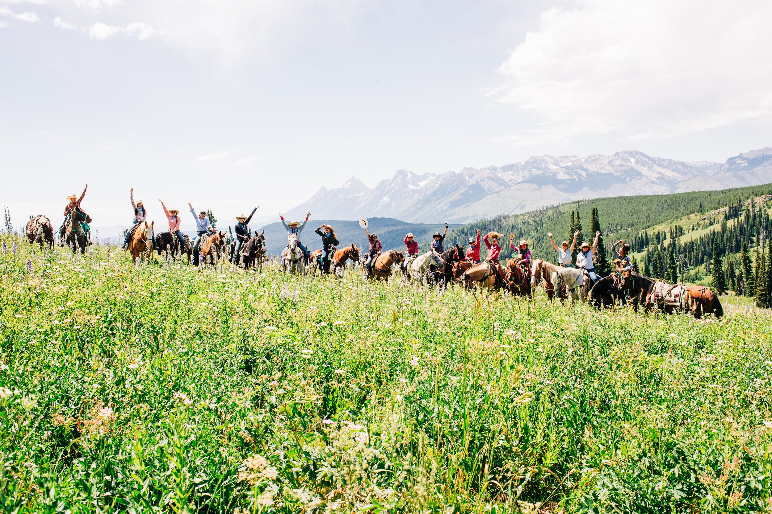 A group of people riding horses through a lush green meadow with mountains in the background. The riders are waving and wearing hats, with some holding hats in the air.