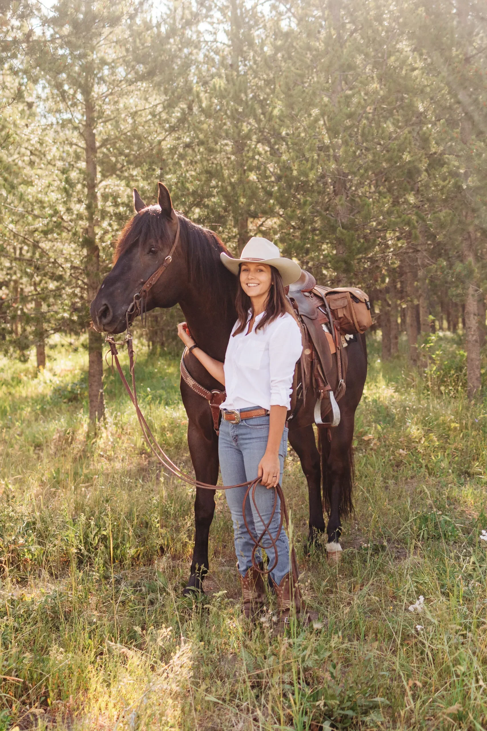 A woman in a white shirt, jeans, cowboy boots, and a cowboy hat standing in a grassy field beside a black horse, with her hand on the horse's neck, in a wooded area with sunlight filtering through the trees.