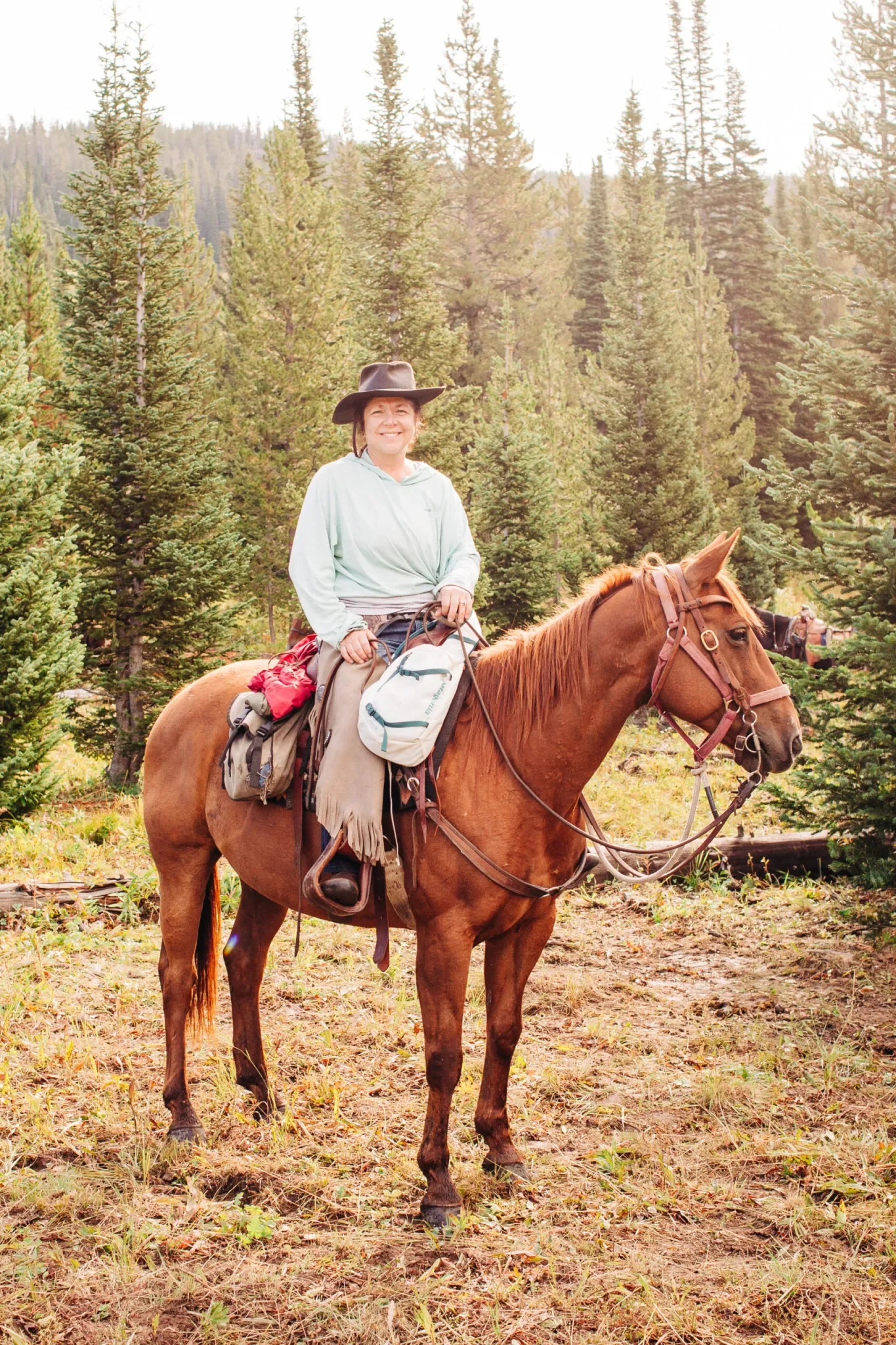 A woman in a light-colored jacket and wide-brimmed hat smiling while sitting on a brown horse in a forest of tall evergreen trees.