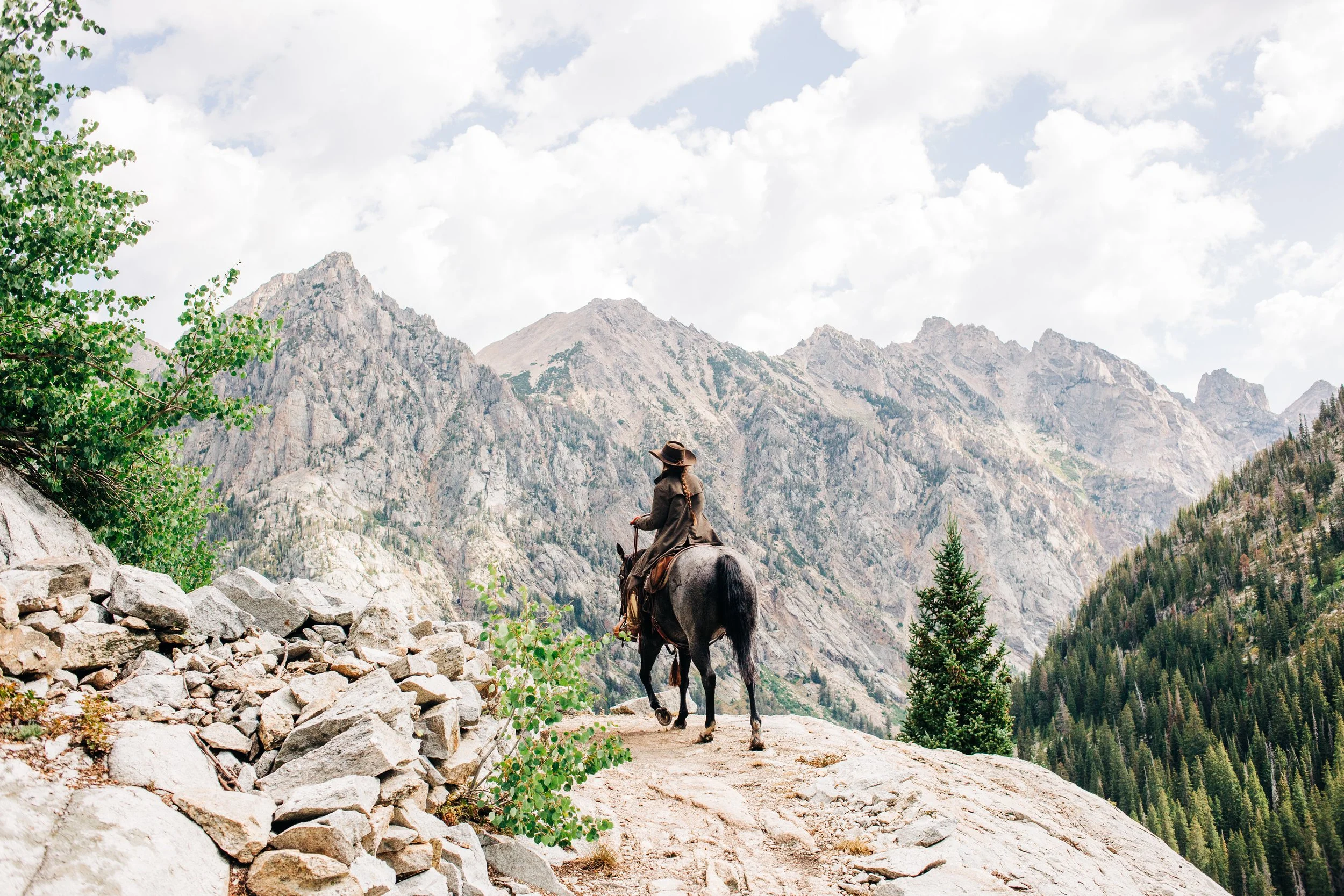 A person riding a horse on a trail with mountain scenery in the background, surrounded by trees and rocks.