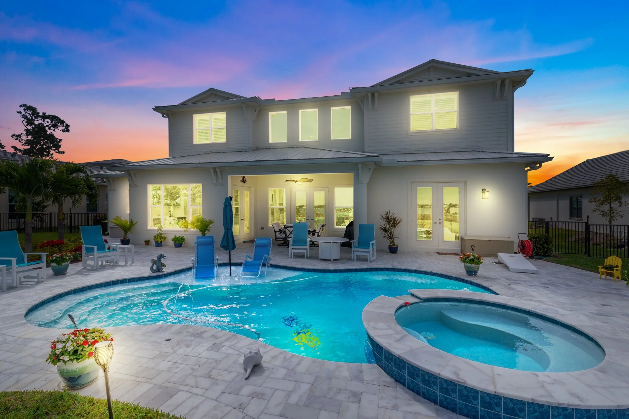 A backyard with a swimming pool, pool chairs, and potted plants in front of a white two-story house with lit windows during sunset.