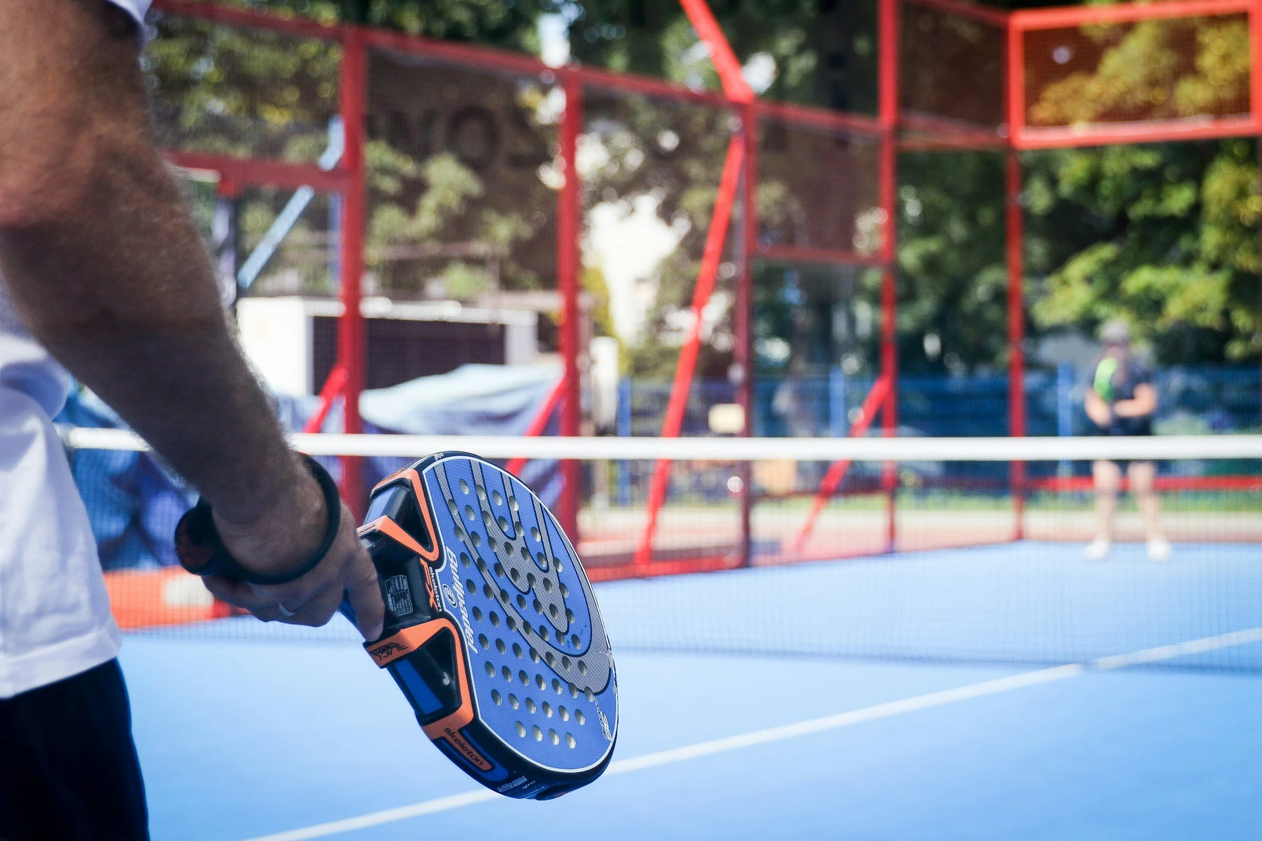 Close-up of a person holding a padel racket on a padel court, with a red fence and trees in the background.