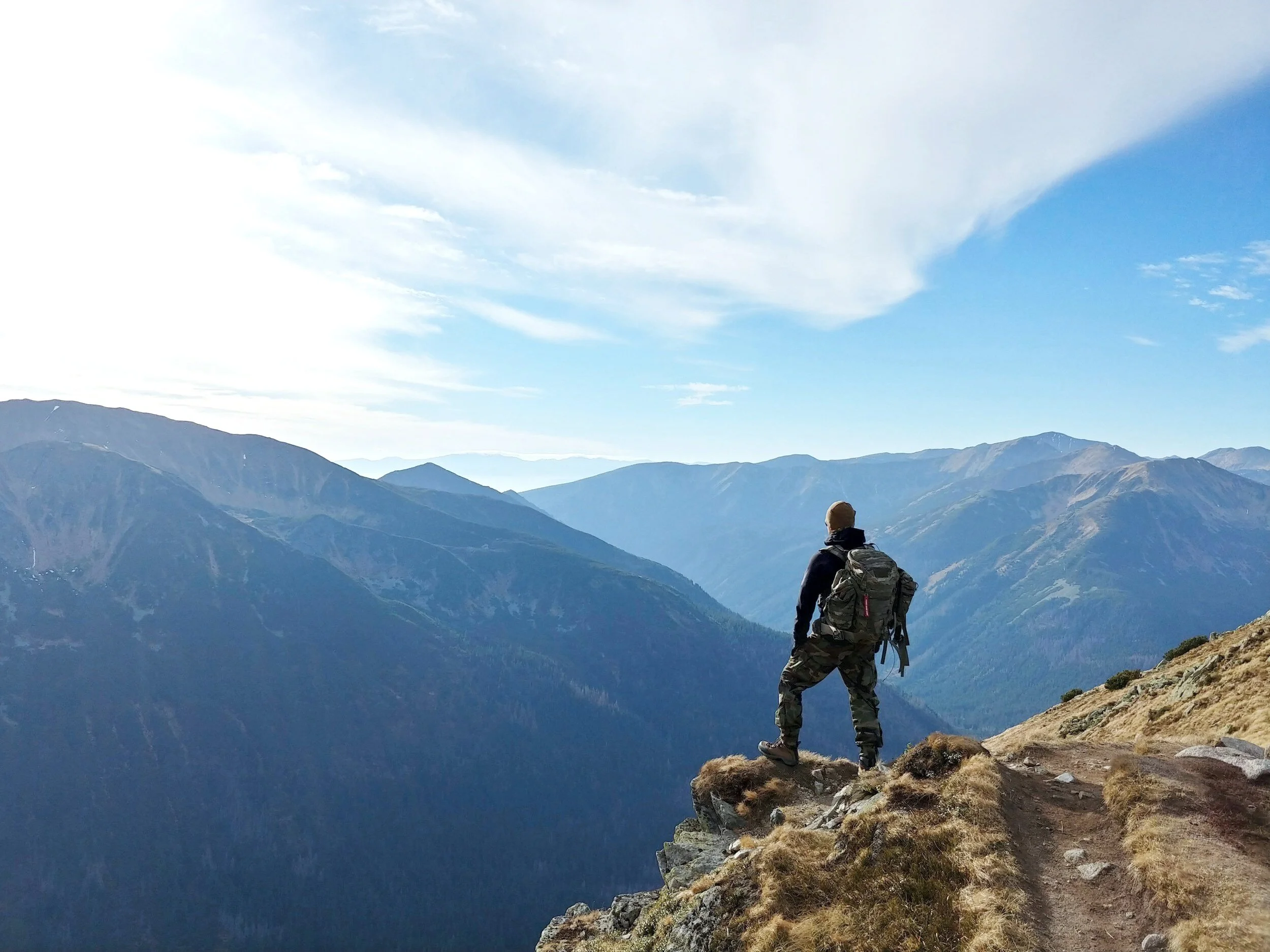A person with a backpack stands on a rocky mountain ledge, overlooking a series of mountain ranges under a partly cloudy sky.