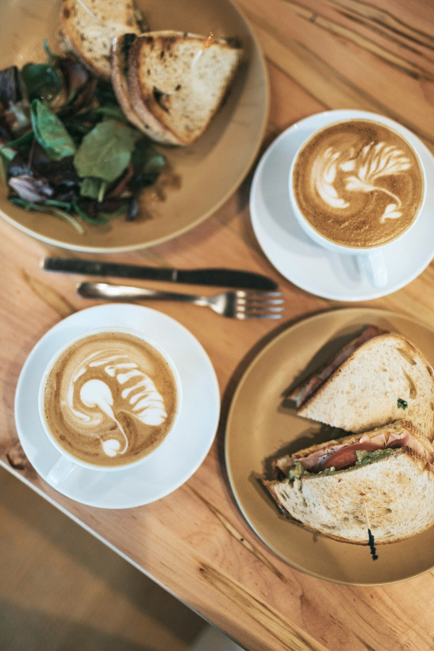 Two cups of coffee with latte art, a plate with toasted bread, and a plate with a sandwich on a wooden table.
