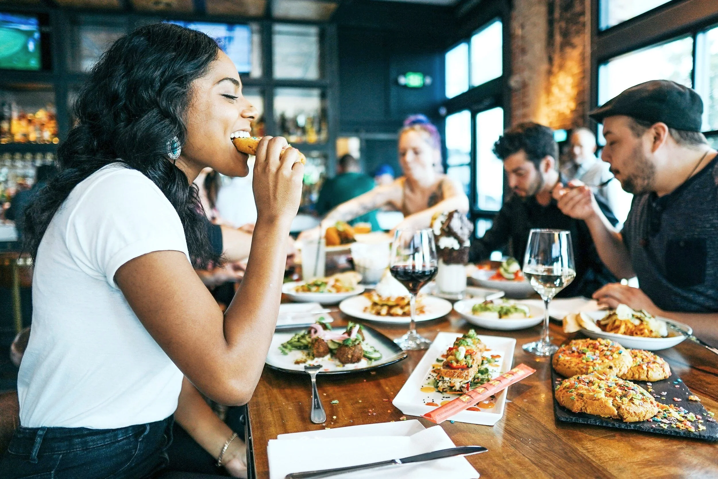 A woman with dark wavy hair wearing a white shirt is eating a fried chicken wing at a restaurant with a group of people seated at a long table filled with various dishes, including desserts and glasses of wine, inside a rustic-modern restaurant with large windows.