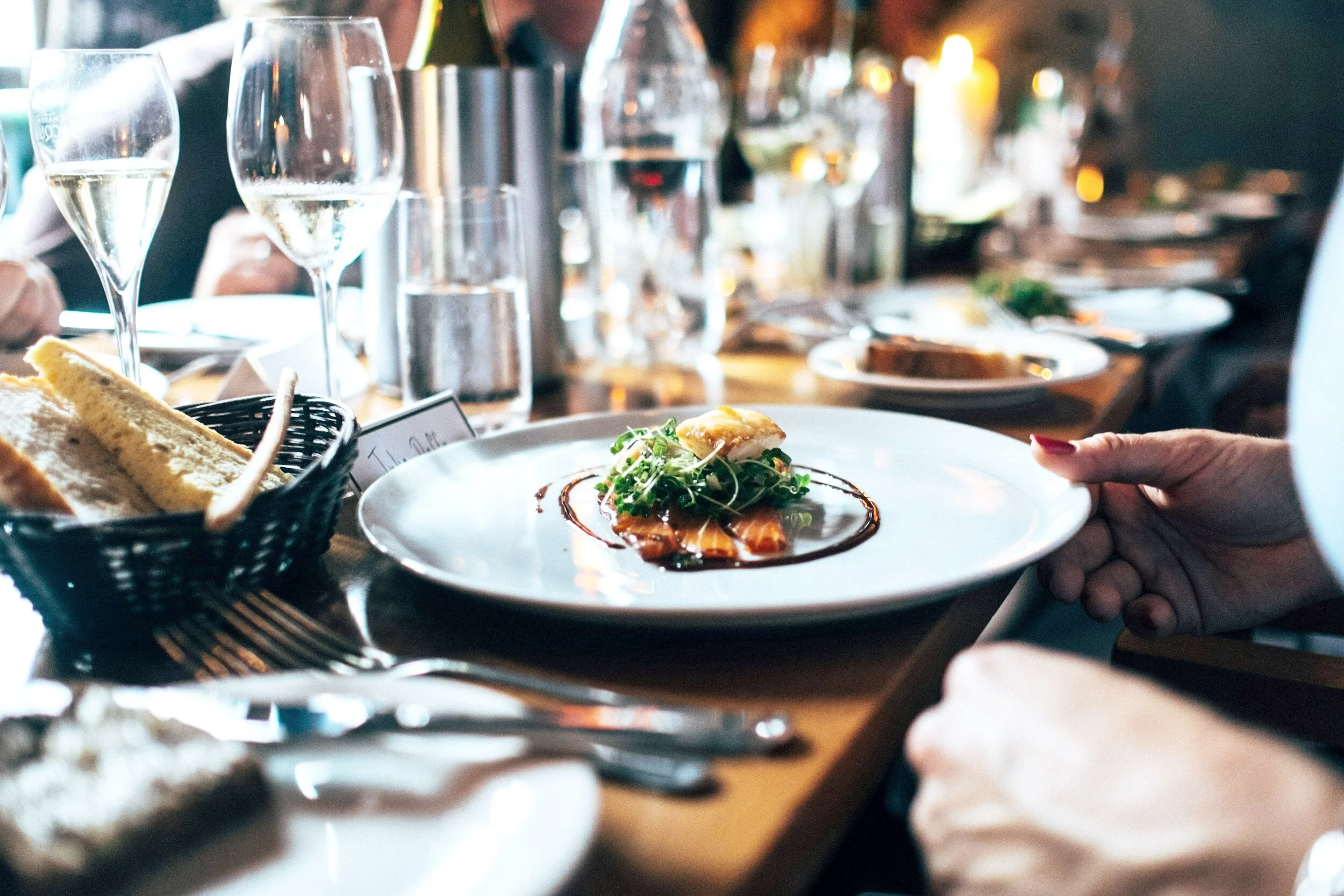 A dinner table at a restaurant with wine glasses, a plate of food with greens and sauce, and bread in a basket. Blurred background with warm lighting.