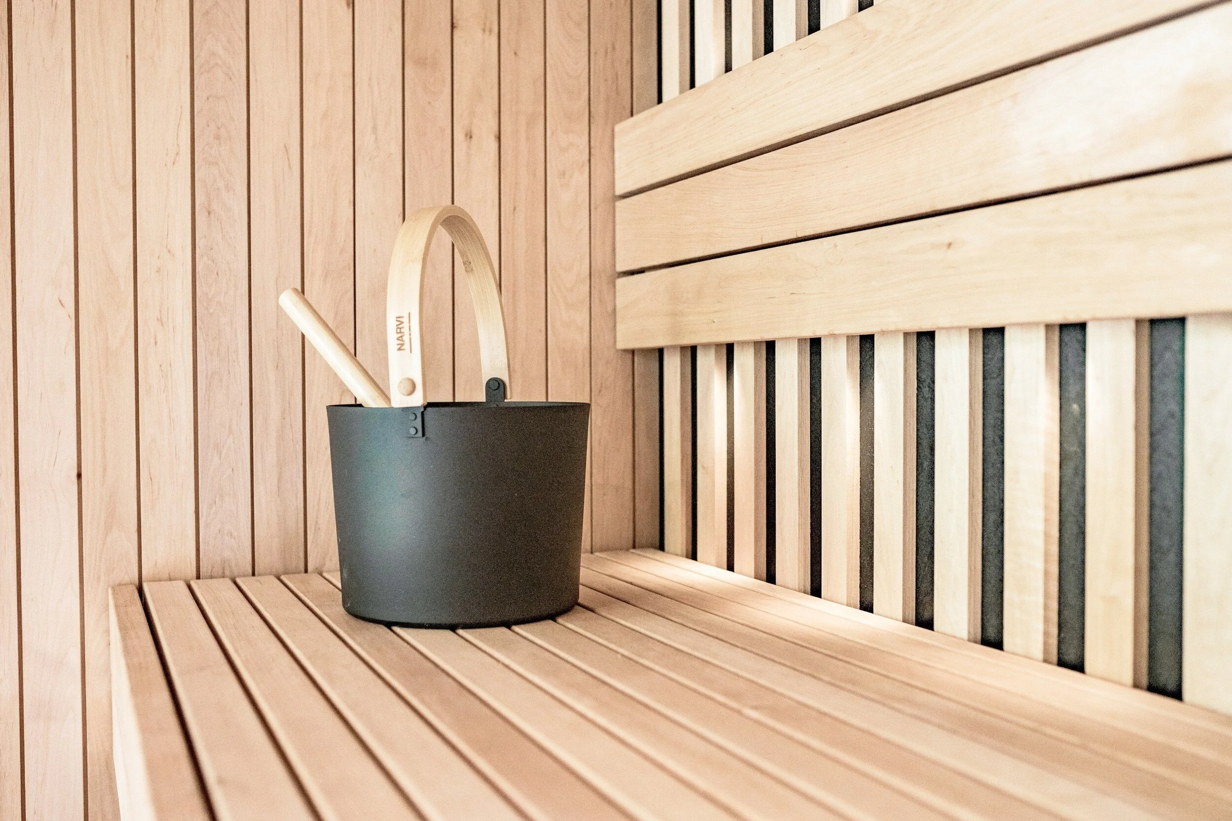 An interior view of a wooden sauna with a black bucket holding a wooden ladle with the word "NARVI" inscribed on it.