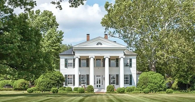 Large white colonial-style house with tall columns, situated on a well-manicured lawn with trees and shrubs.