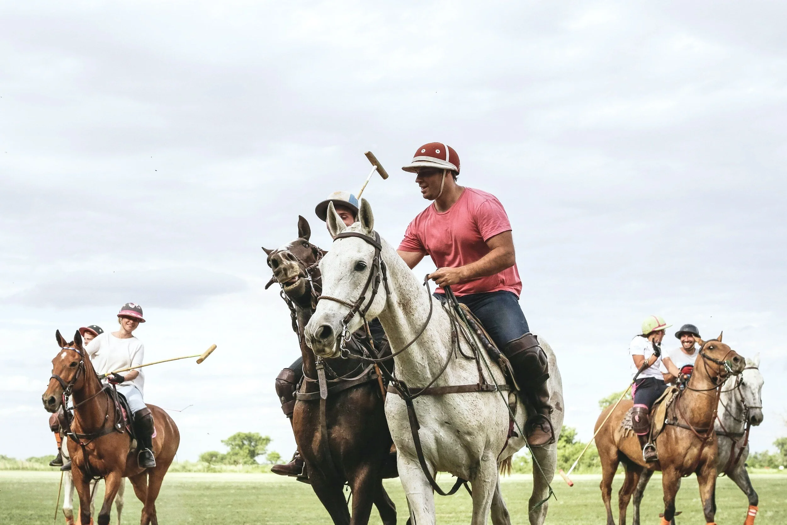 Group of people riding horses on a grassy field, some wearing helmets, against a cloudy sky.