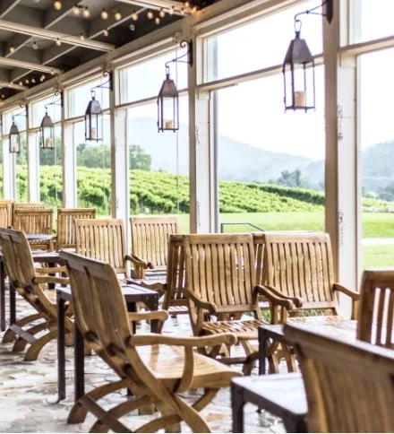Outdoor patio seating with wooden chairs and tables, lantern-style light fixtures, large windows, and scenic green landscape view.