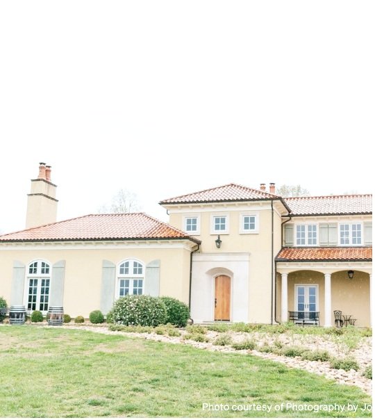 A large, two-story house with a beige exterior, red-tiled roof, and multiple windows surrounded by a well-maintained lawn and landscaping.
