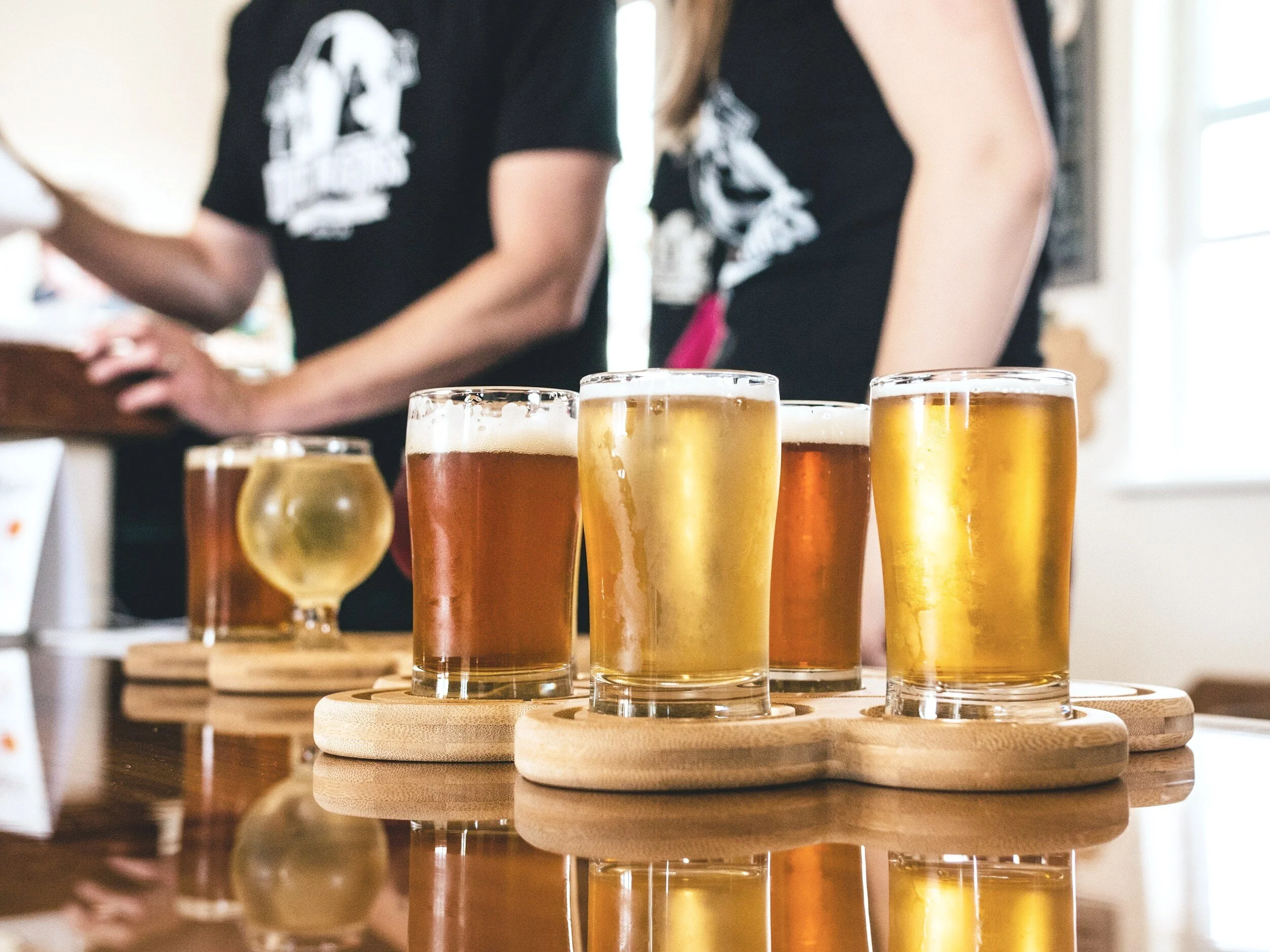 Several glasses of beer of different types and colors on round wooden coasters, set on a wooden table, with a blur of people in the background.