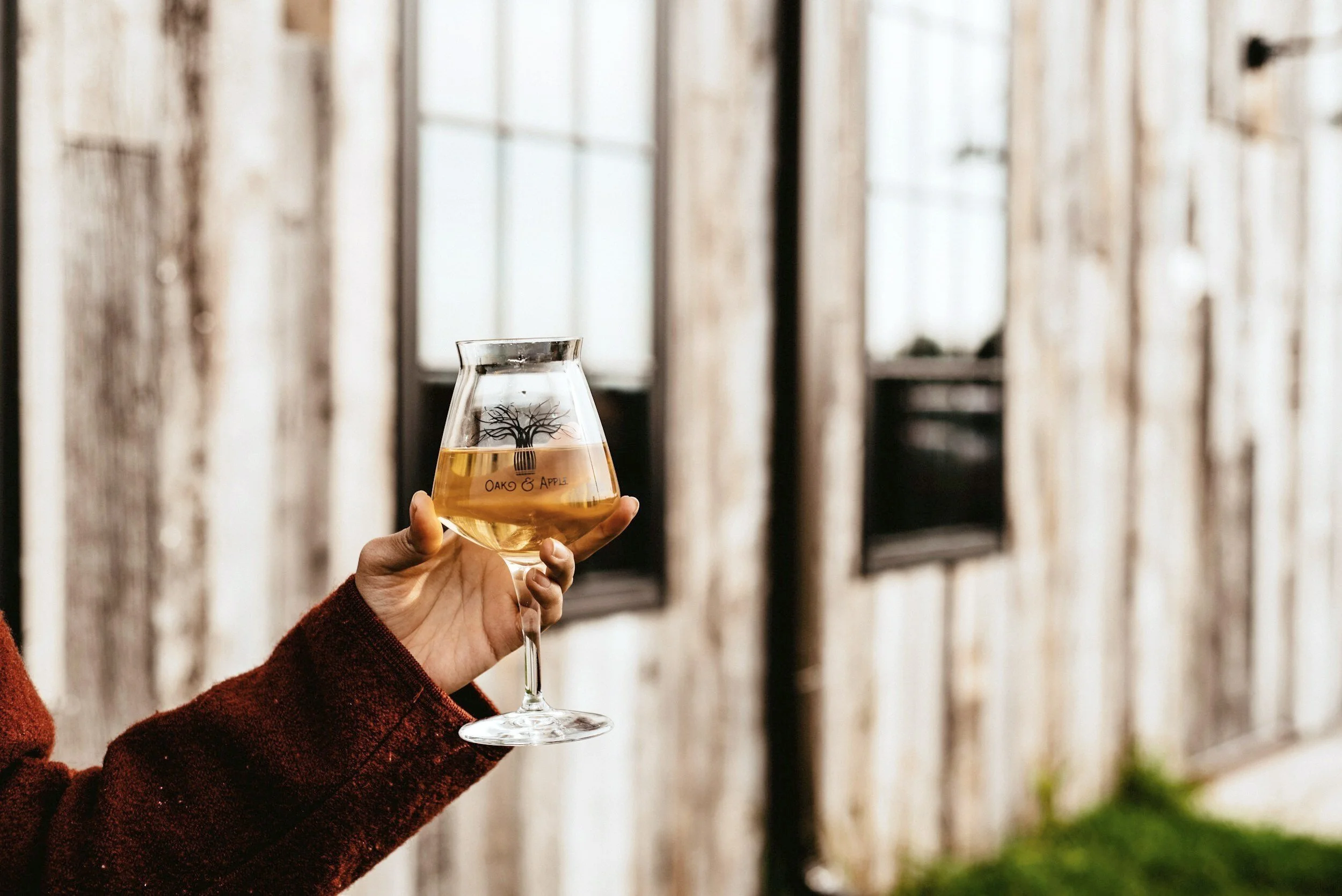 A hand holding a glass of white wine with a tree logo and the text 'OAK & APPLE' on it, in front of a wooden fence.