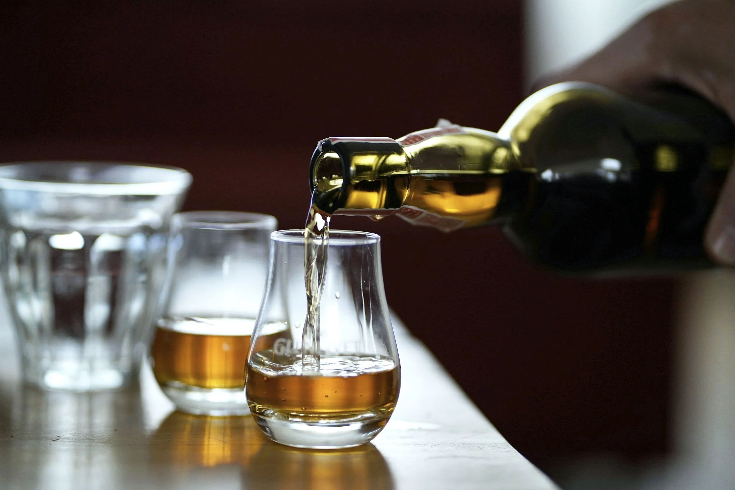 A person pouring a beverage from a green bottle into a glass on a wooden surface, with several empty glasses nearby.