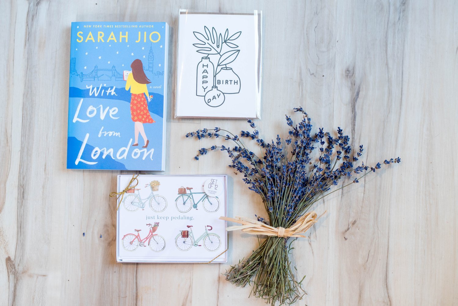 A flat lay of a bouquet of dried lavender tied with a straw ribbon, a cartoon card with bicycle illustrations and the phrase 'just keep pedaling,' a birthday card with floral drawings, a book titled 'With Love from London' by Sarah Jio, and a small greeting card with a minimalist design and the words 'HAPPY BIRTHDAY' on a light wooden surface.