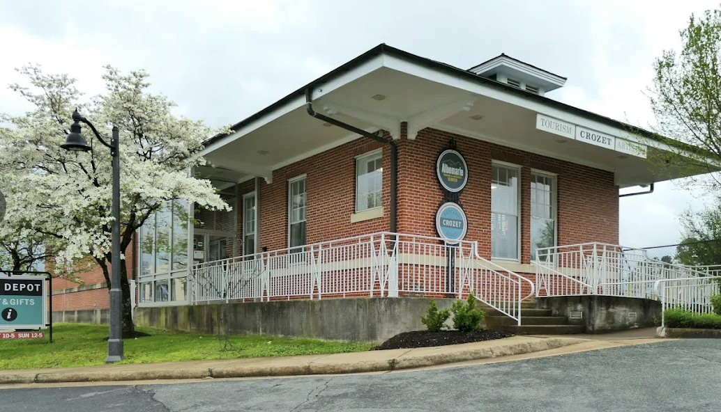 A red brick building with a white overhang, large windows, and signs indicating it is a tourism and arts information center, with a sidewalk, small plants, a streetlamp, blossoming trees, and a parking lot in the foreground.